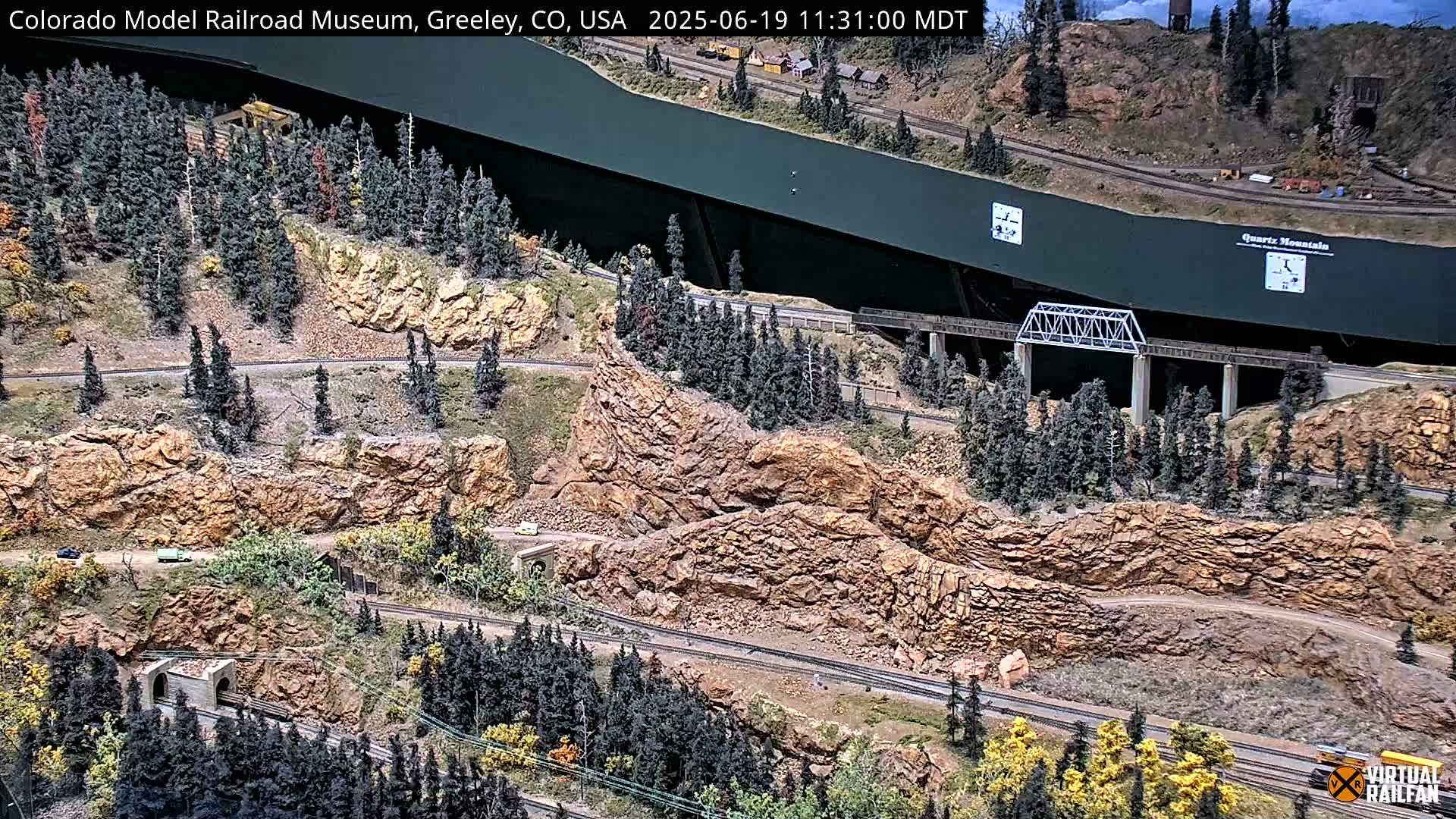 A model railroad display depicts a mountainous landscape with winding roads, tunnels, a bridge, and small vehicles, under a sunny sky.