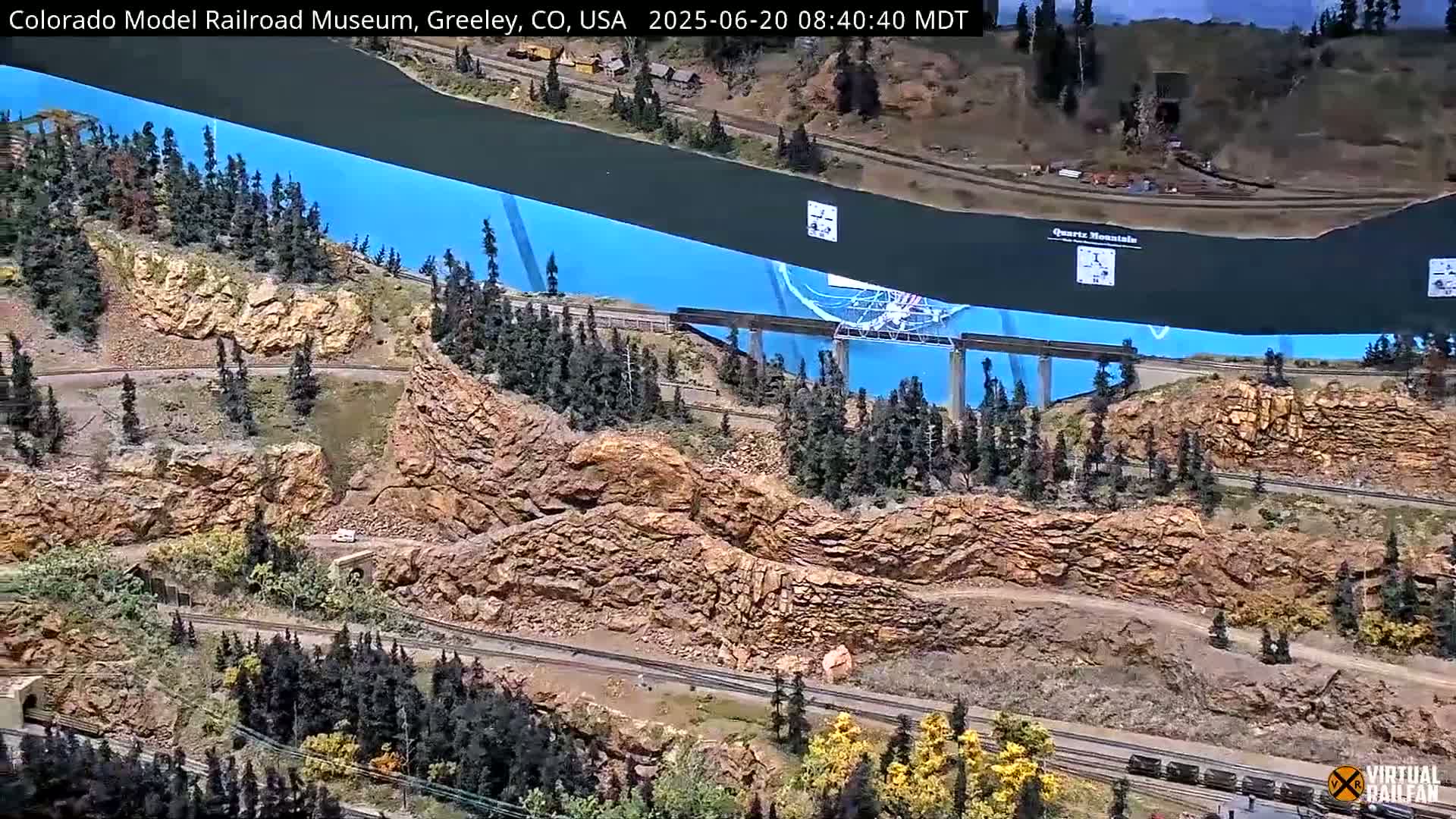 An expansive model railroad landscape features miniature mountains, winding roads, and train tracks, with a large blue backdrop.