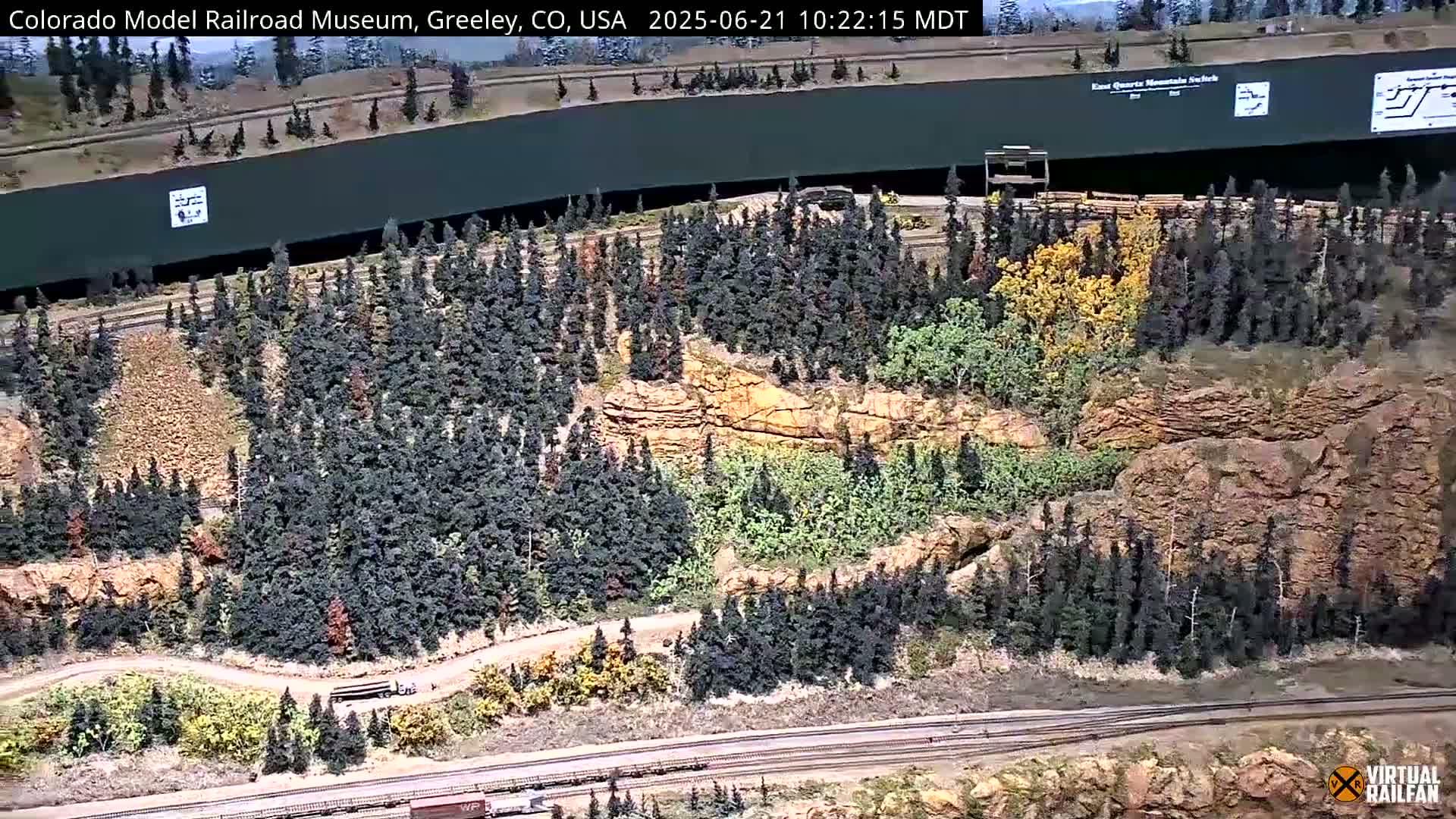 An aerial view of a large model train set depicting a mountainous landscape with coniferous and deciduous trees, a rocky cliff face, and a long, dark structure possibly a tunnel or building, under a sunny sky.