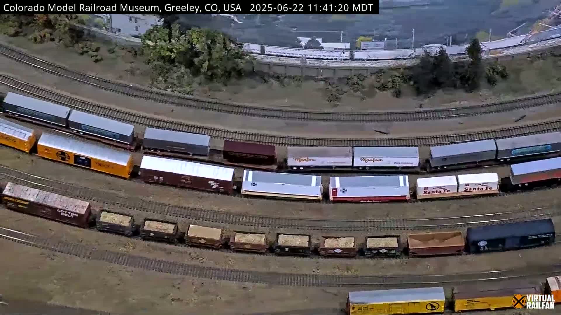 An aerial view shows a model train curving around a track, with various boxcars and hopper cars in different colors and markings.