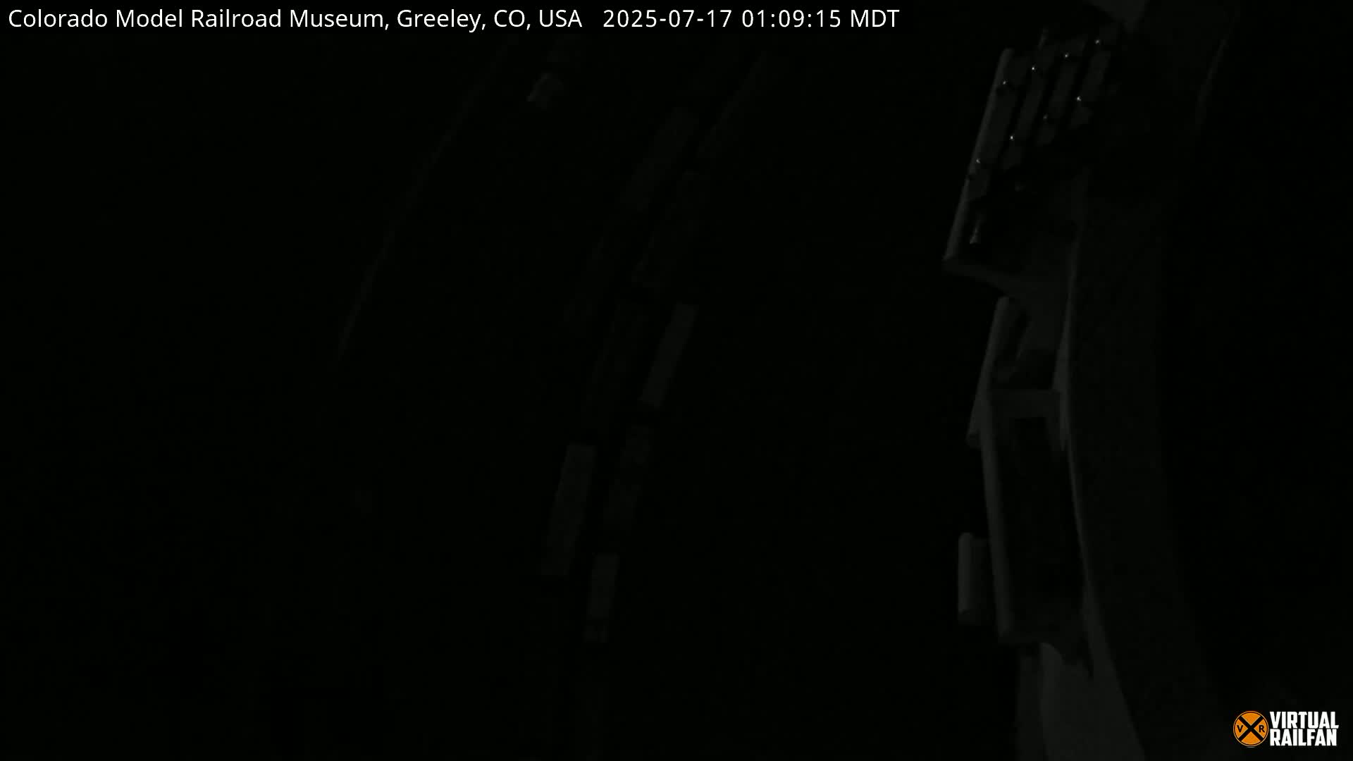 A dark, nighttime image shows a portion of a model railroad track and structures.