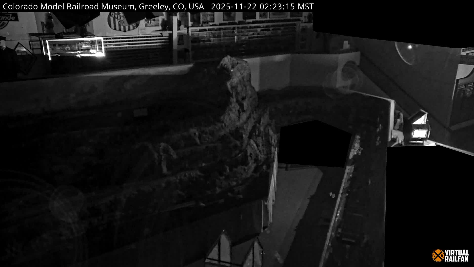 A low-light, black and white image displays an elevated view of a model railroad layout, featuring a large rock formation, miniature buildings, and various illuminated scenes.