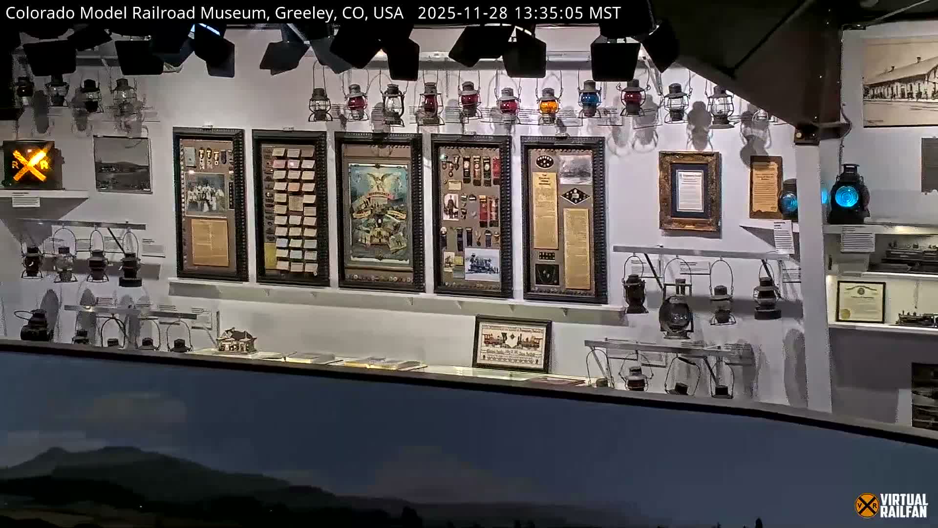 An indoor museum exhibit features a wall covered with framed historical documents, photographs, and collections of small objects, alongside various railroad lanterns, situated above a detailed model landscape in the foreground.