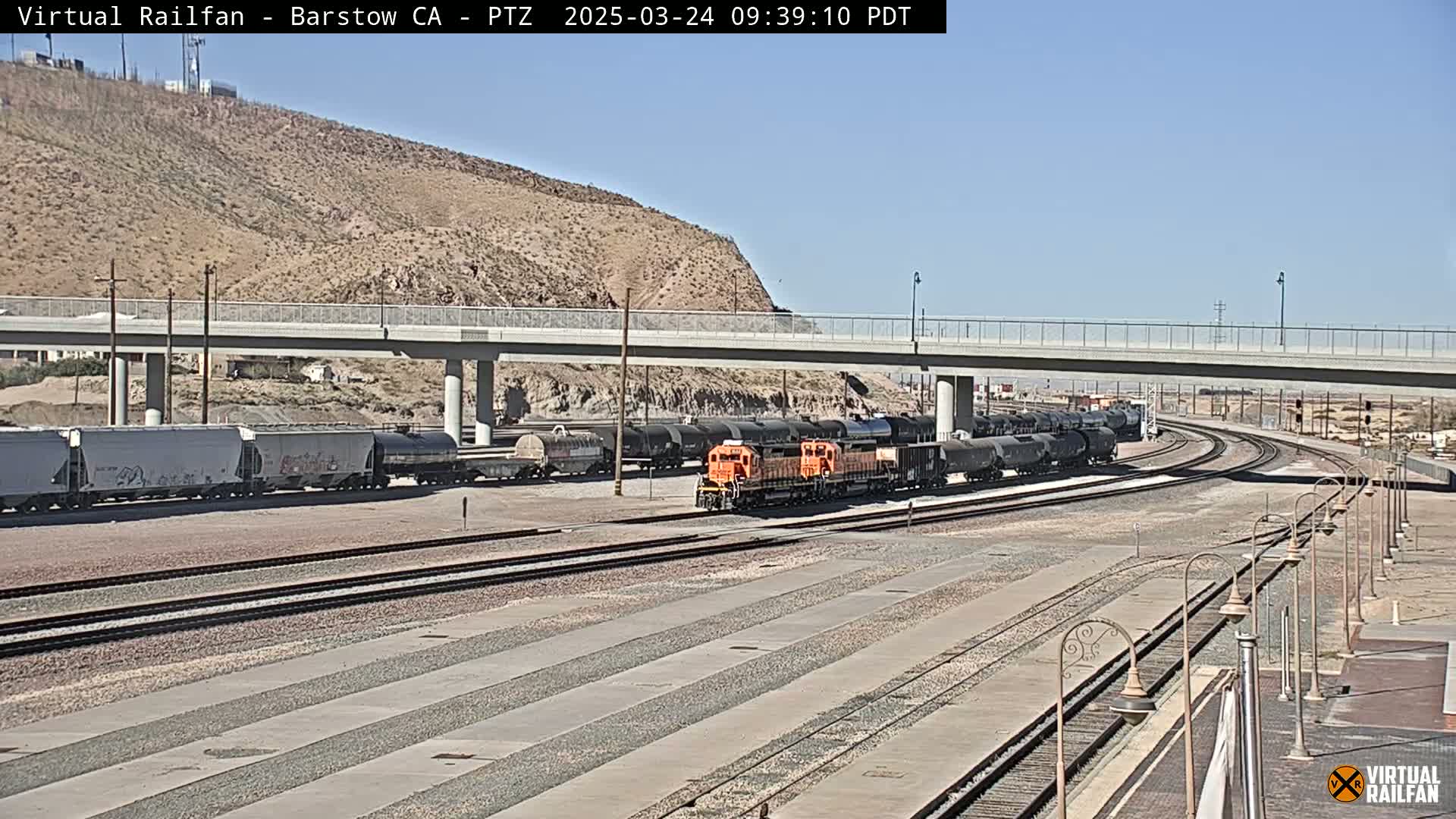 An orange locomotive leads a long train of dark tanker cars on a rail yard under a clear sky near a hill and overpass.