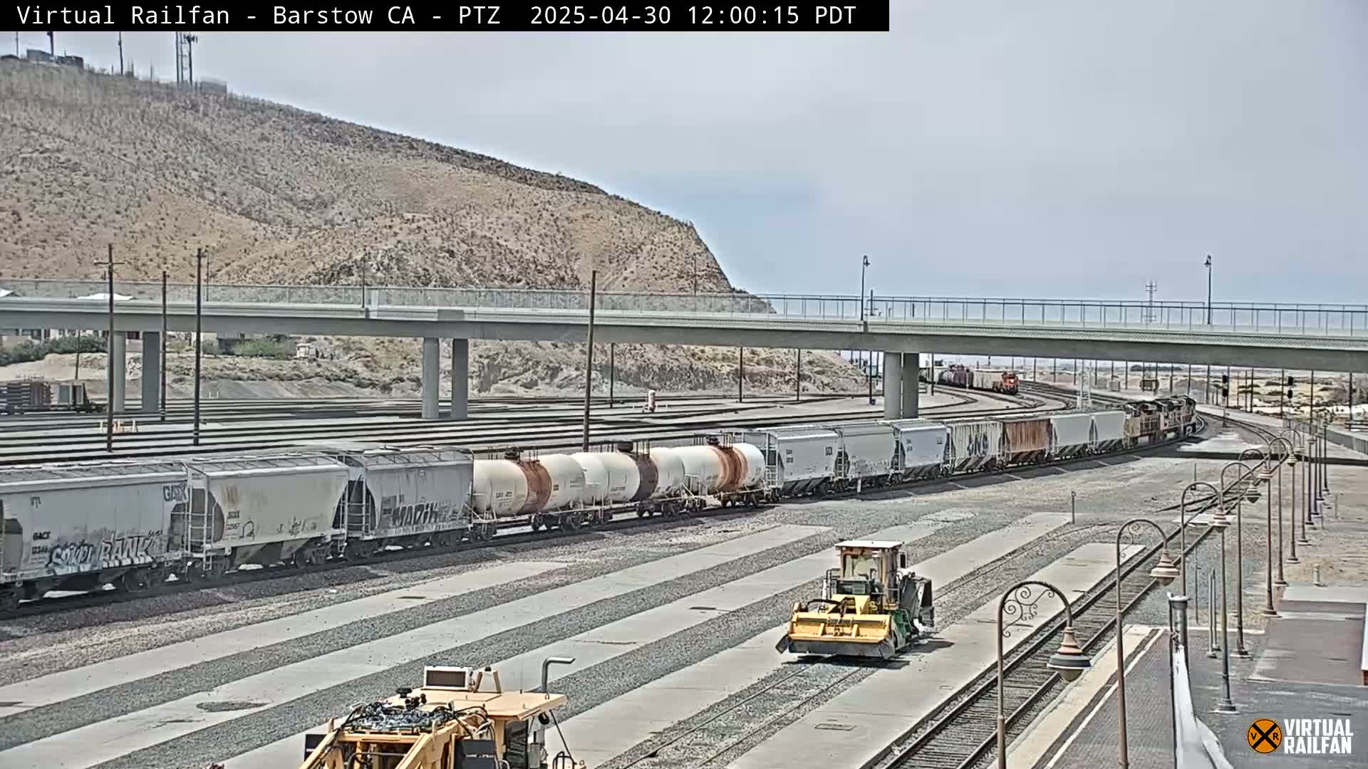 A long freight train sits in a rail yard under a partly sunny sky, with a road grader and other construction equipment nearby.
