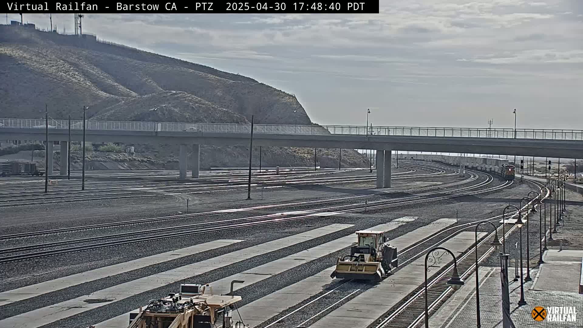 A long freight train passes under a bridge on a mostly clear day, with some light construction equipment visible near train tracks.