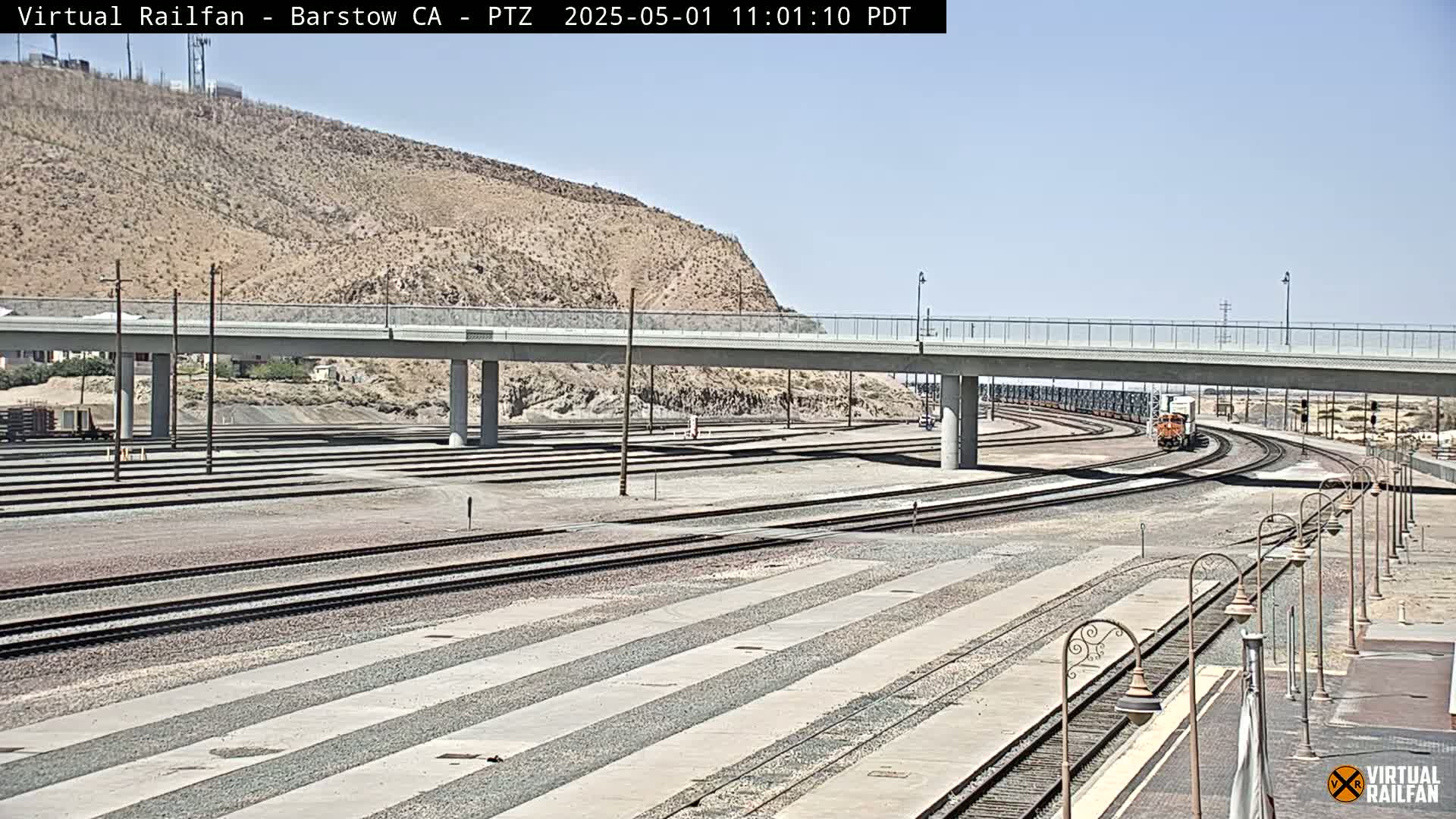 A train passes under a bridge on a sunny day near a sprawling rail yard.