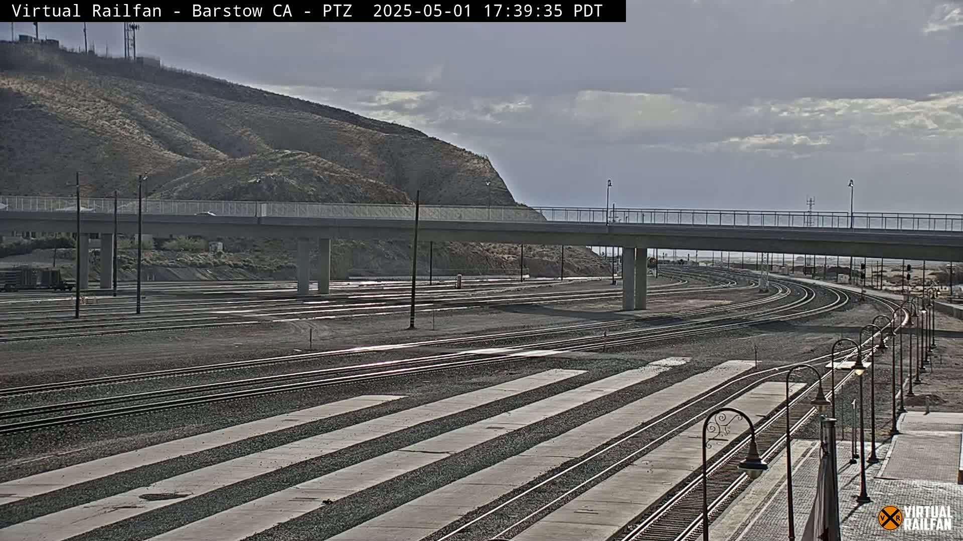 A wide view of a railway yard under a cloudy sky, featuring multiple train tracks leading to a highway overpass against a backdrop of a brown hill.