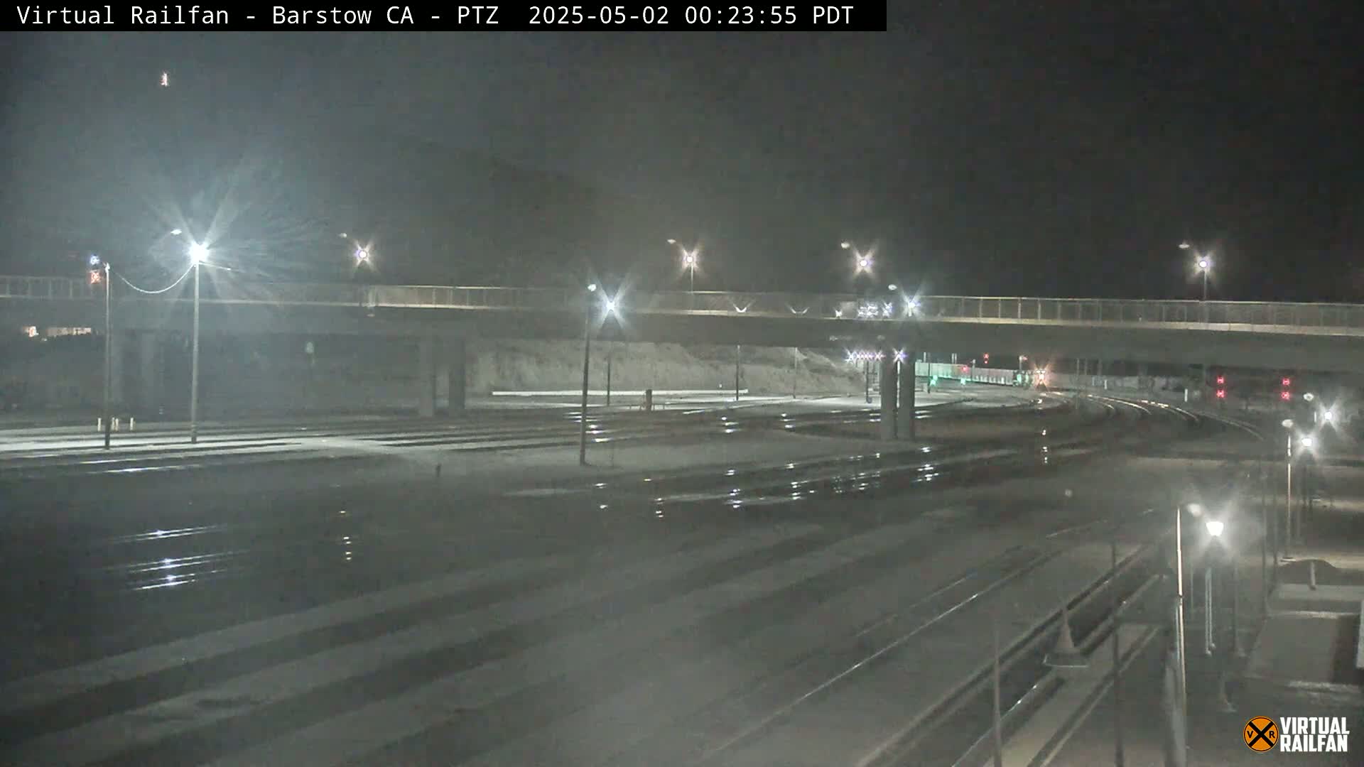 A nighttime view of a snow-covered train yard with a road overpass under bright lights.