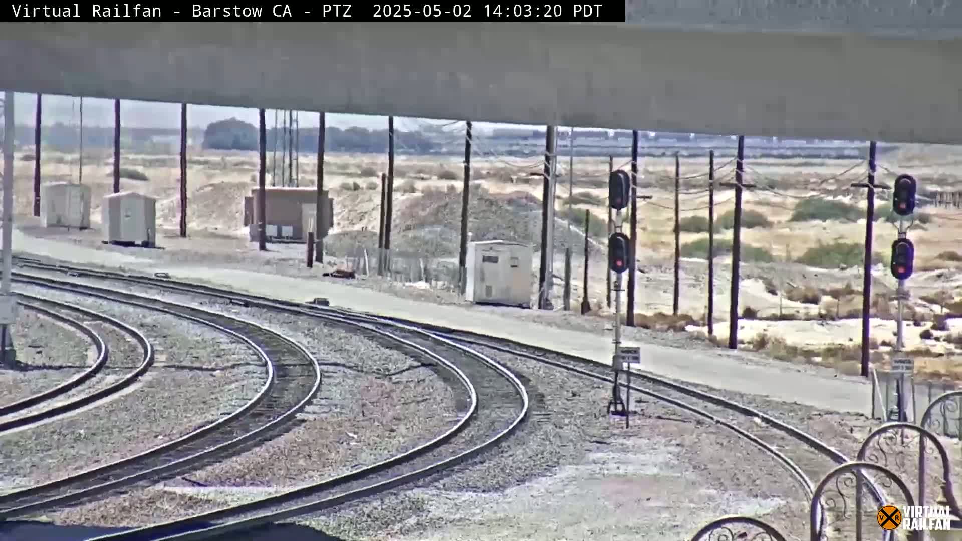 A sunny outdoor view shows several curving train tracks next to a roadway, with utility boxes and signal lights visible in a dry, desert-like landscape.