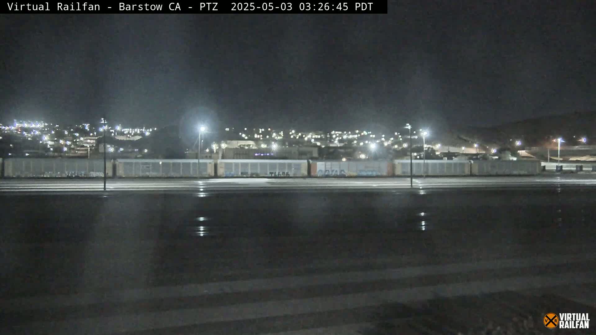 A nighttime view of a rail yard with a town in the background, under slightly wet conditions.
