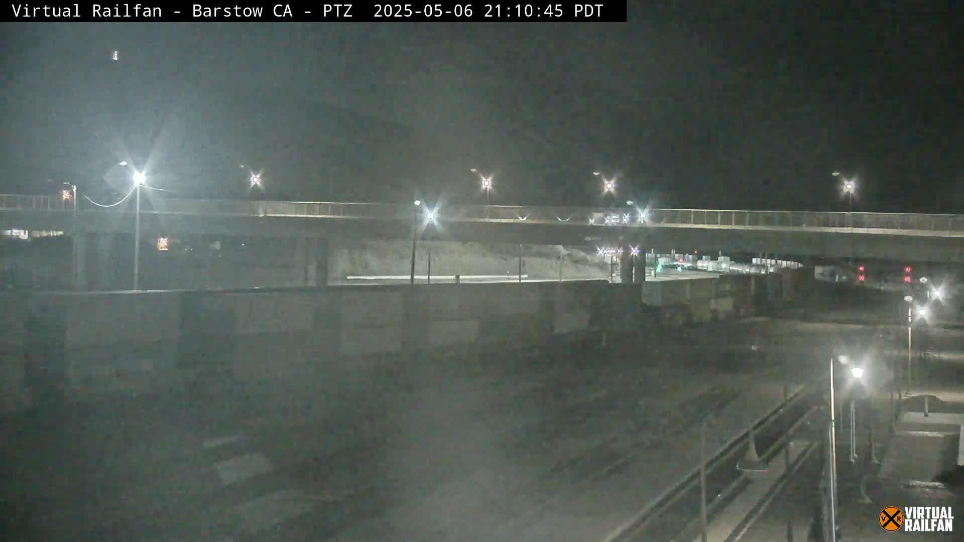 A nighttime view of a train yard under a bridge, illuminated by streetlights.