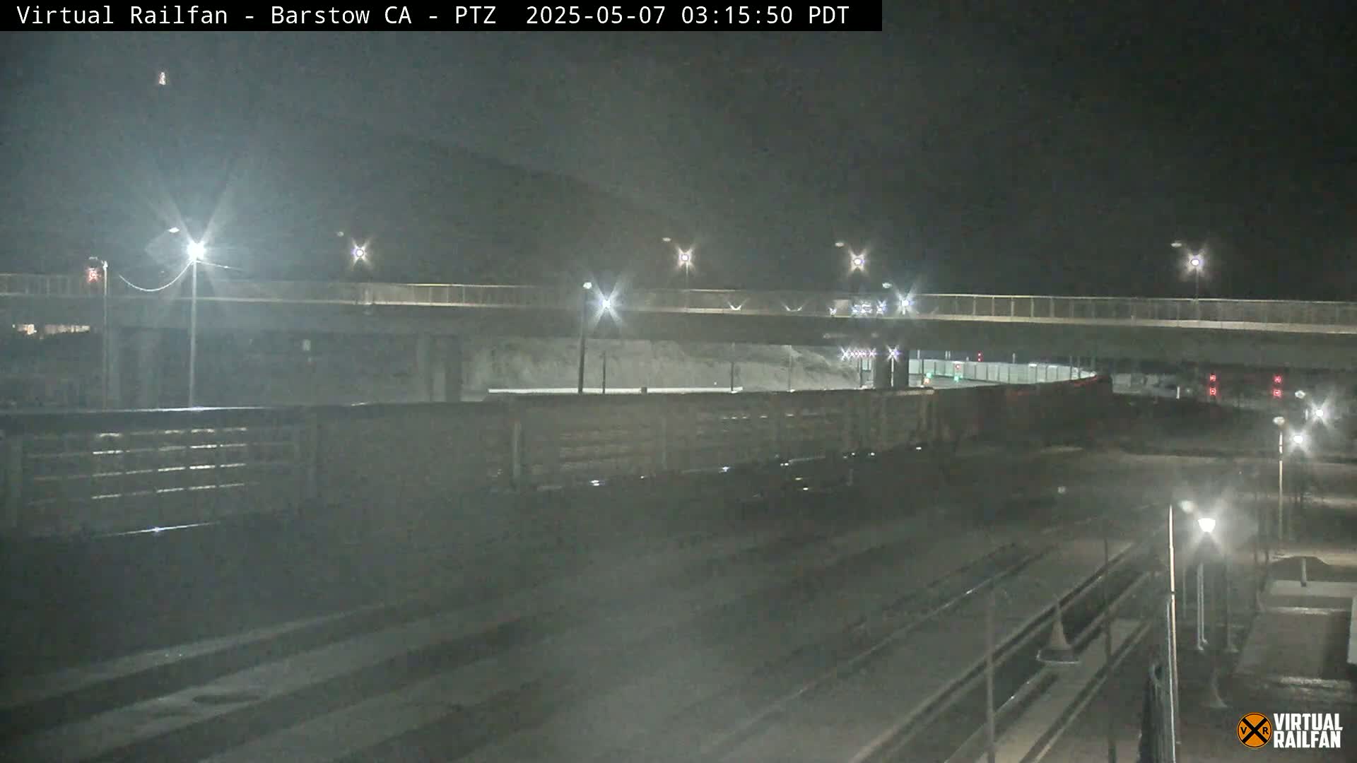 A long freight train sits at night under a highway overpass, illuminated by streetlights.