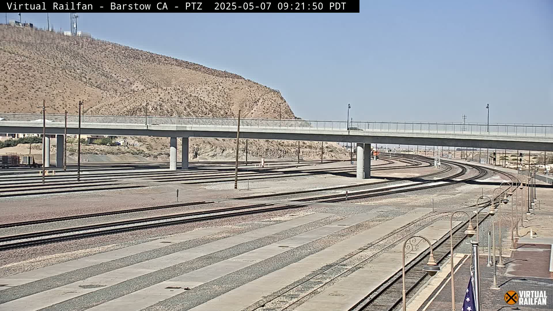 A wide shot of a railway yard under a clear sky, showing numerous train tracks that curve around a concrete overpass, with a hill visible in the background.