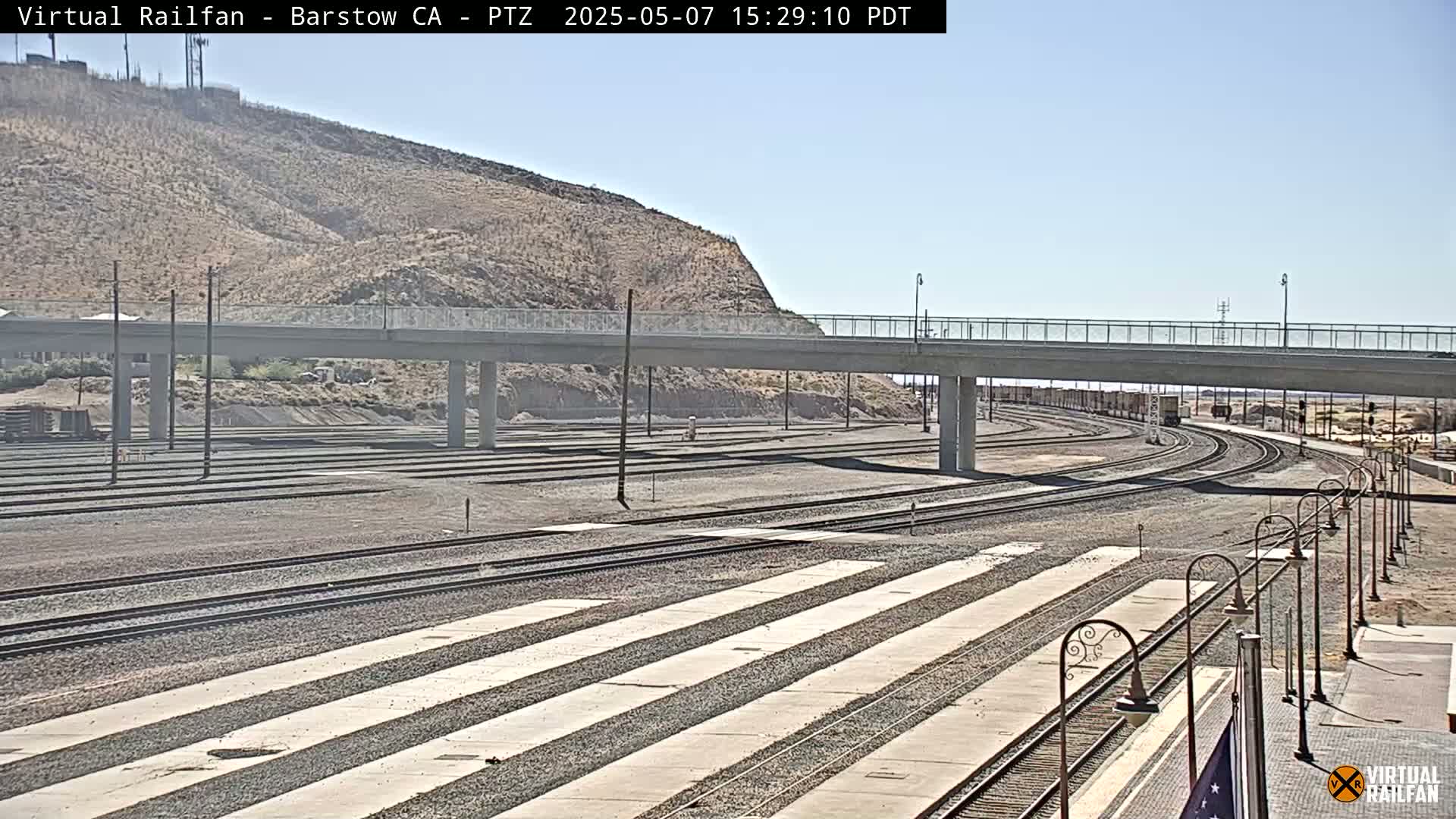 A sunny day reveals a train yard with numerous tracks, a concrete overpass, and a hill in the background.