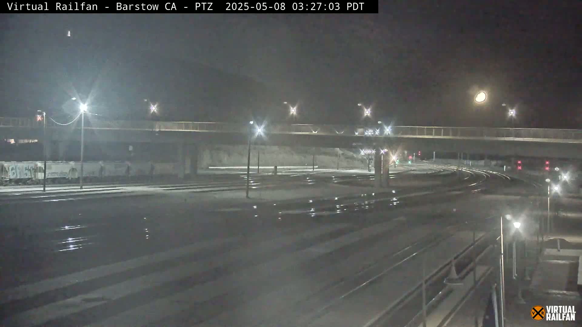 A nighttime view of a snow-covered train yard with a bridge overhead and a full moon visible in the sky.