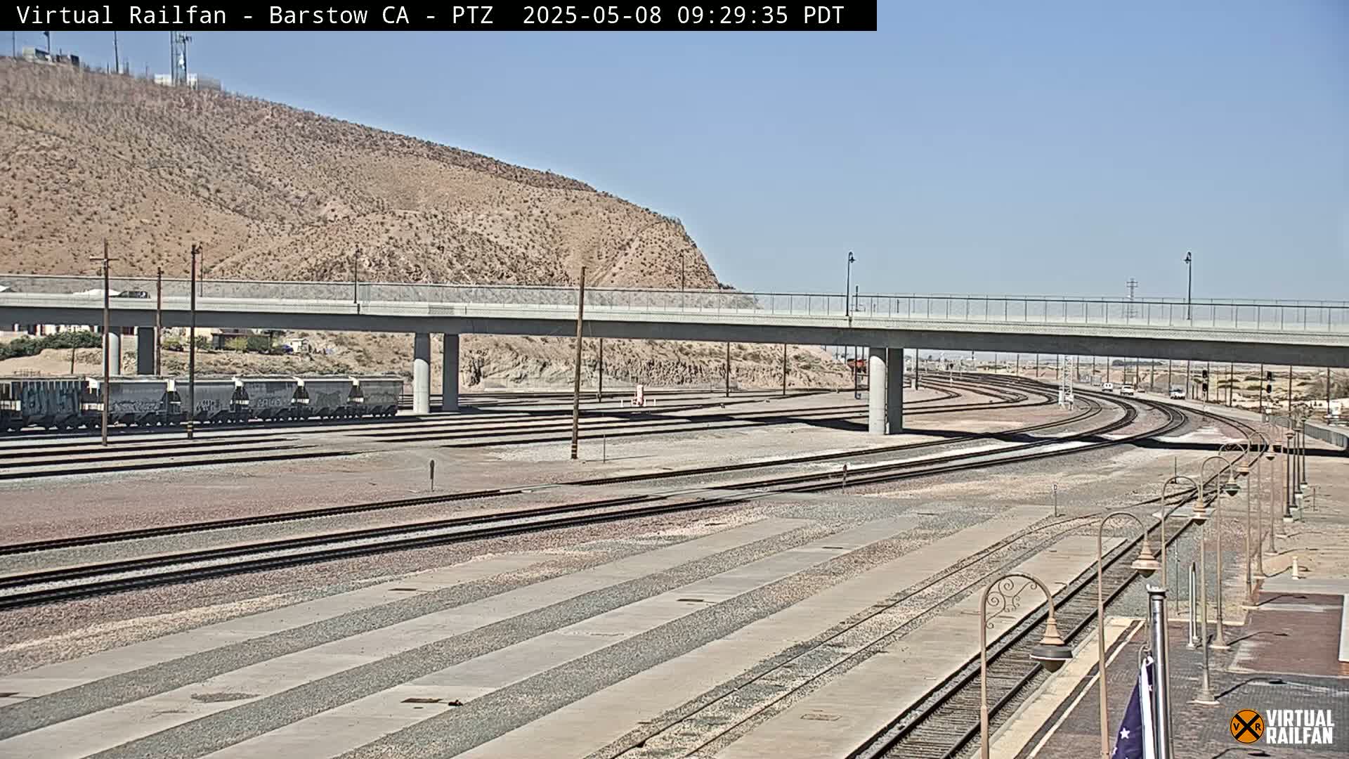 A train yard with several sets of railroad tracks under a clear sky, next to a highway overpass and a desert hillside.