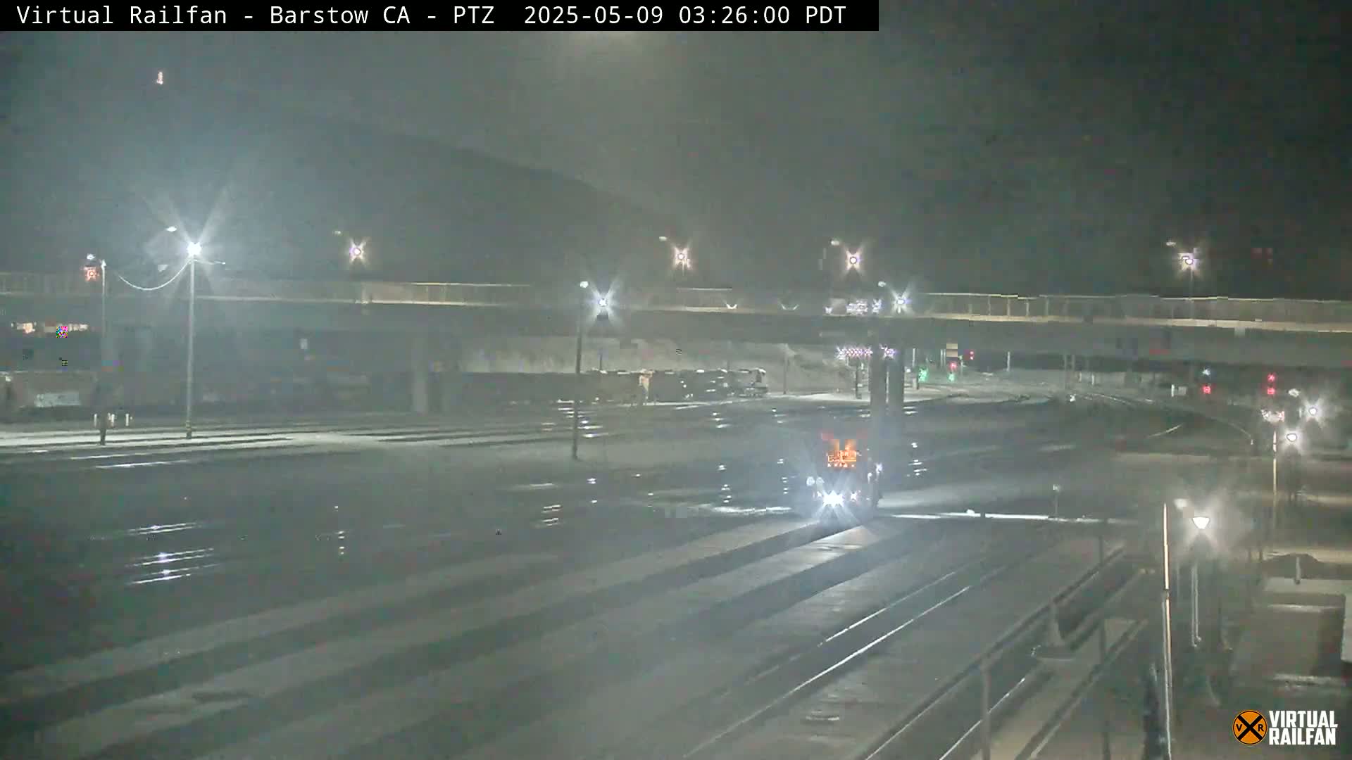 A nighttime view of train tracks and a roadway under a bridge, with a vehicle visible on the tracks.