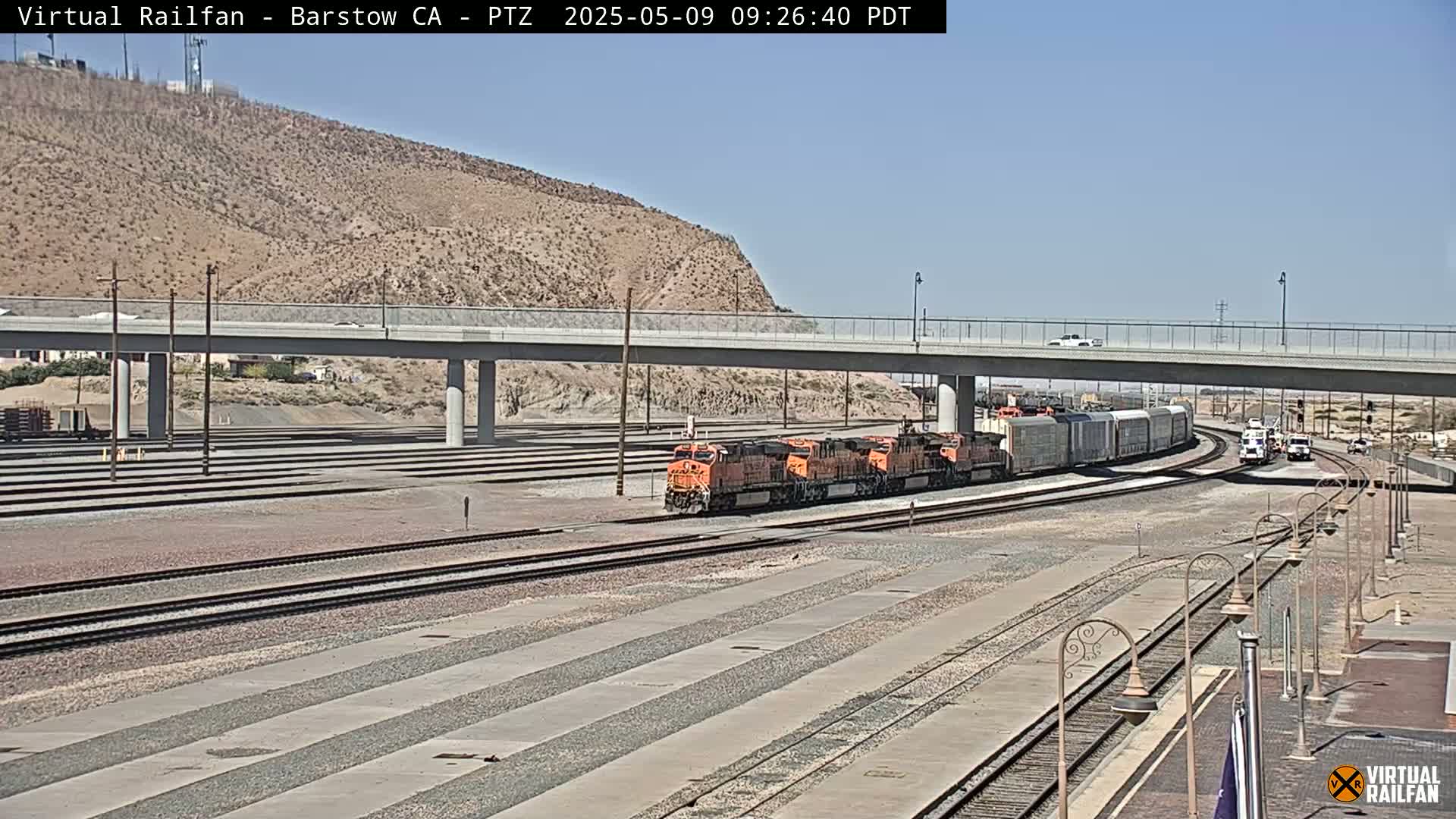 A freight train and several orange locomotives are on a railway track under a bridge on a sunny day, with a barren hill in the background.