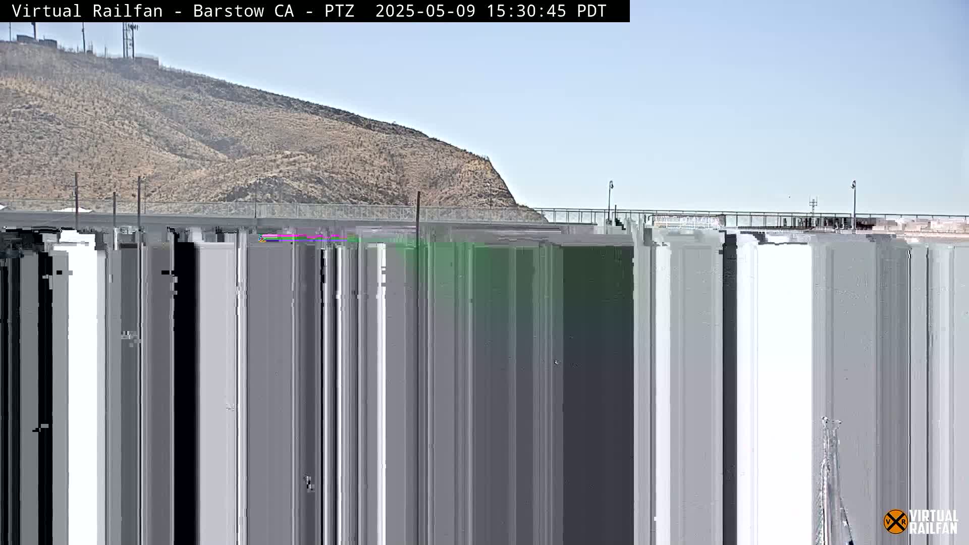 A mostly clear sky is over a tan hill behind a bridge and a long structure of tall, grey, vertical columns.