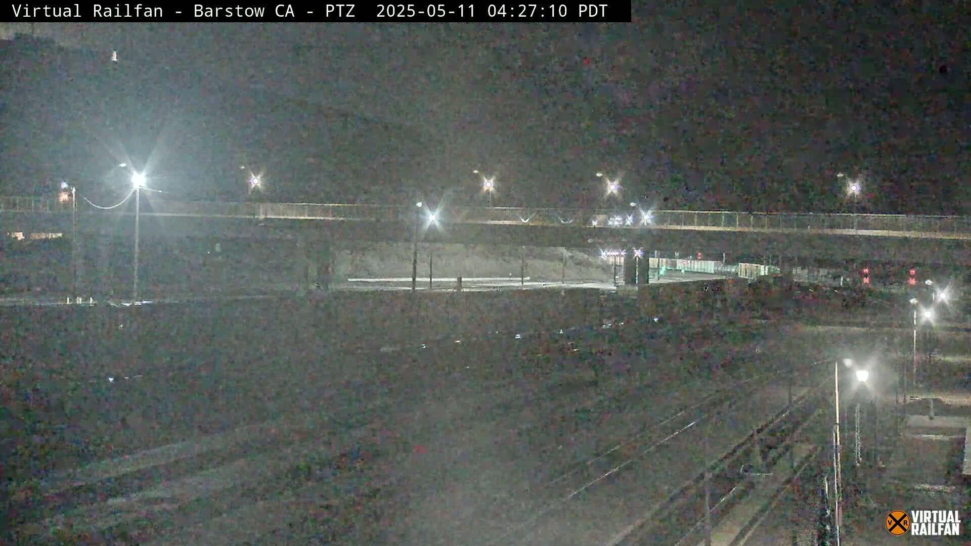 A nighttime view of a train track below a highway overpass, illuminated by streetlights.