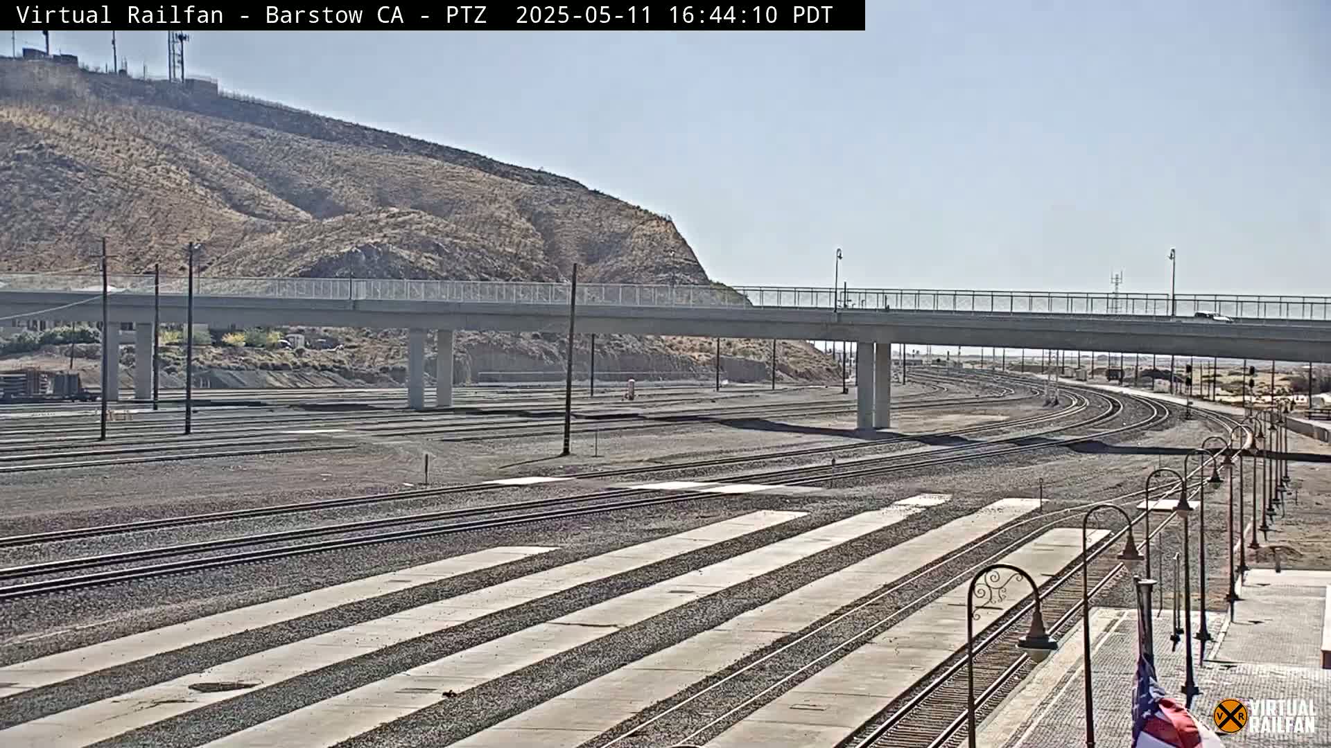 A wide shot of railroad tracks and a bridge under a clear, sunny sky, with a hill visible in the background.
