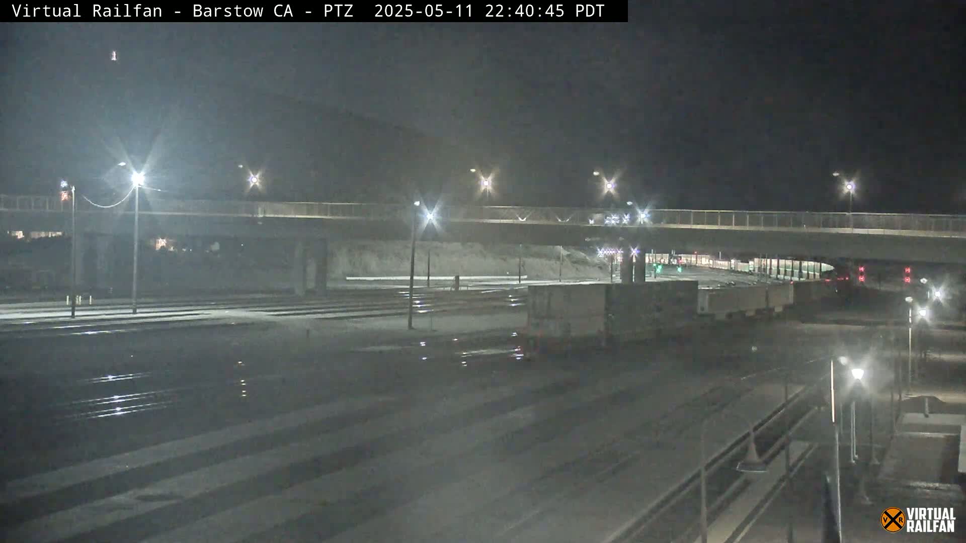 A nighttime view of a snow-covered train yard with a road overpass, lit by streetlights, and a freight train partially visible.