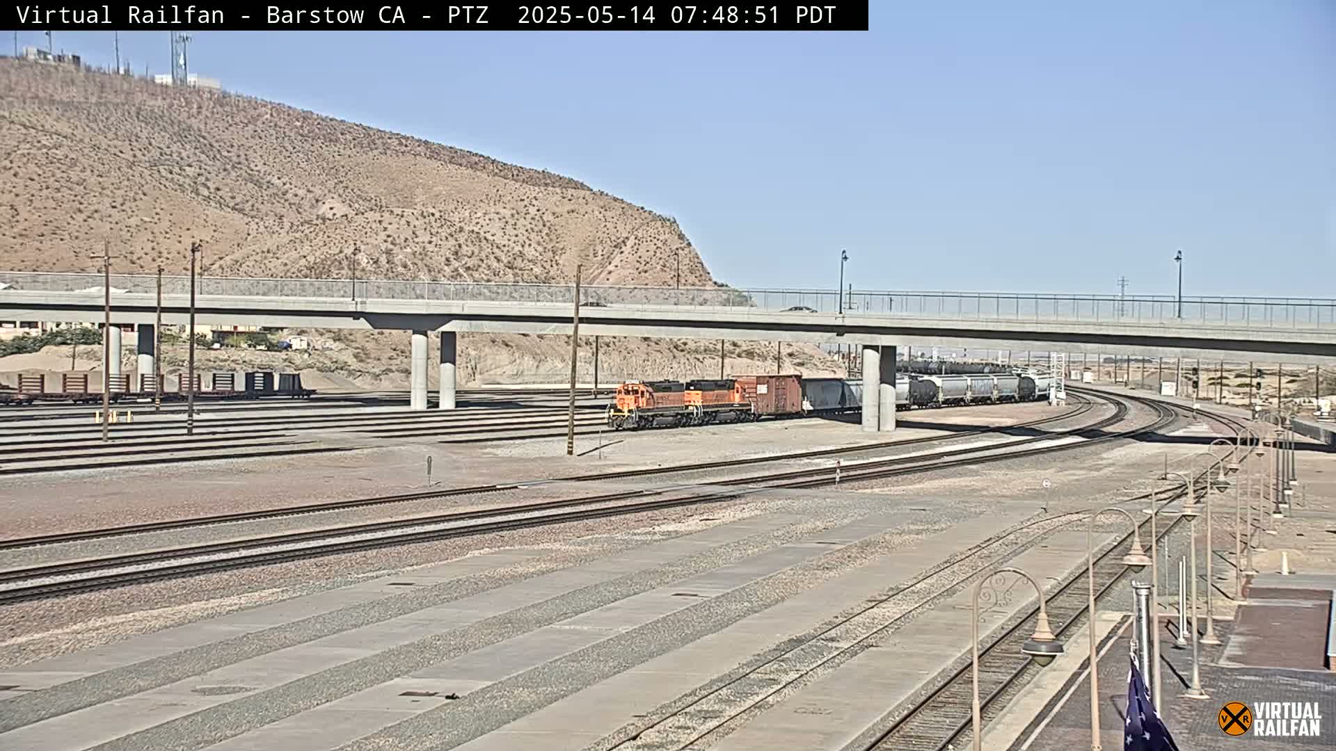A freight train sits on a set of railroad tracks beneath a highway overpass on a sunny day.