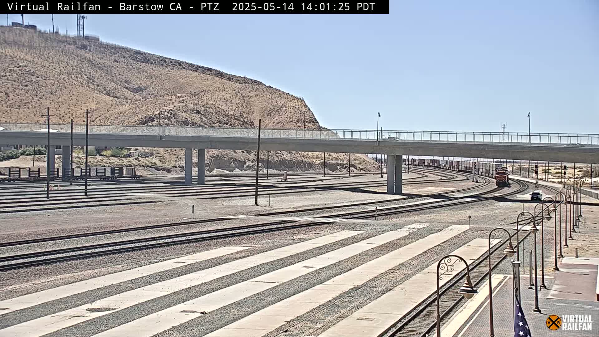 A freight train travels along a network of railroad tracks under a bridge on a sunny day, with a dry, hilly landscape in the background.