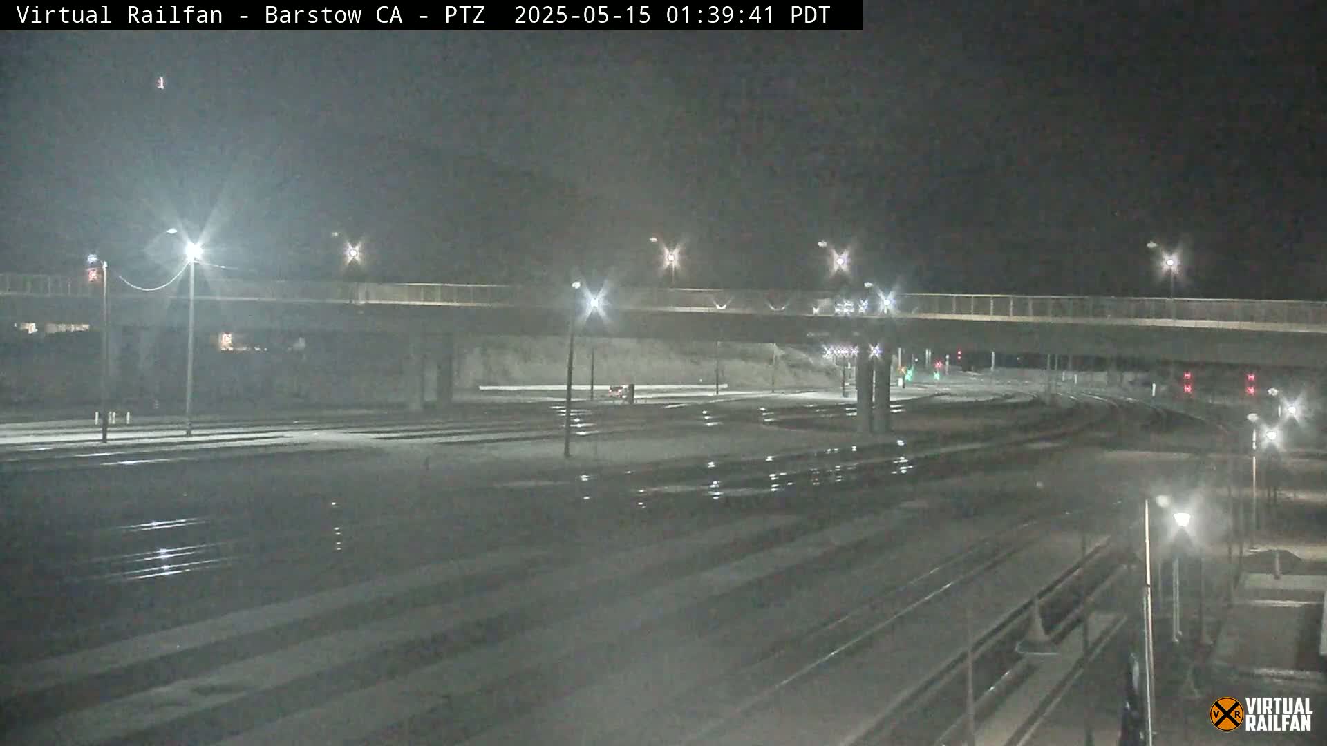 A nighttime, snowy scene shows train tracks and an overpass with lights illuminating the area.