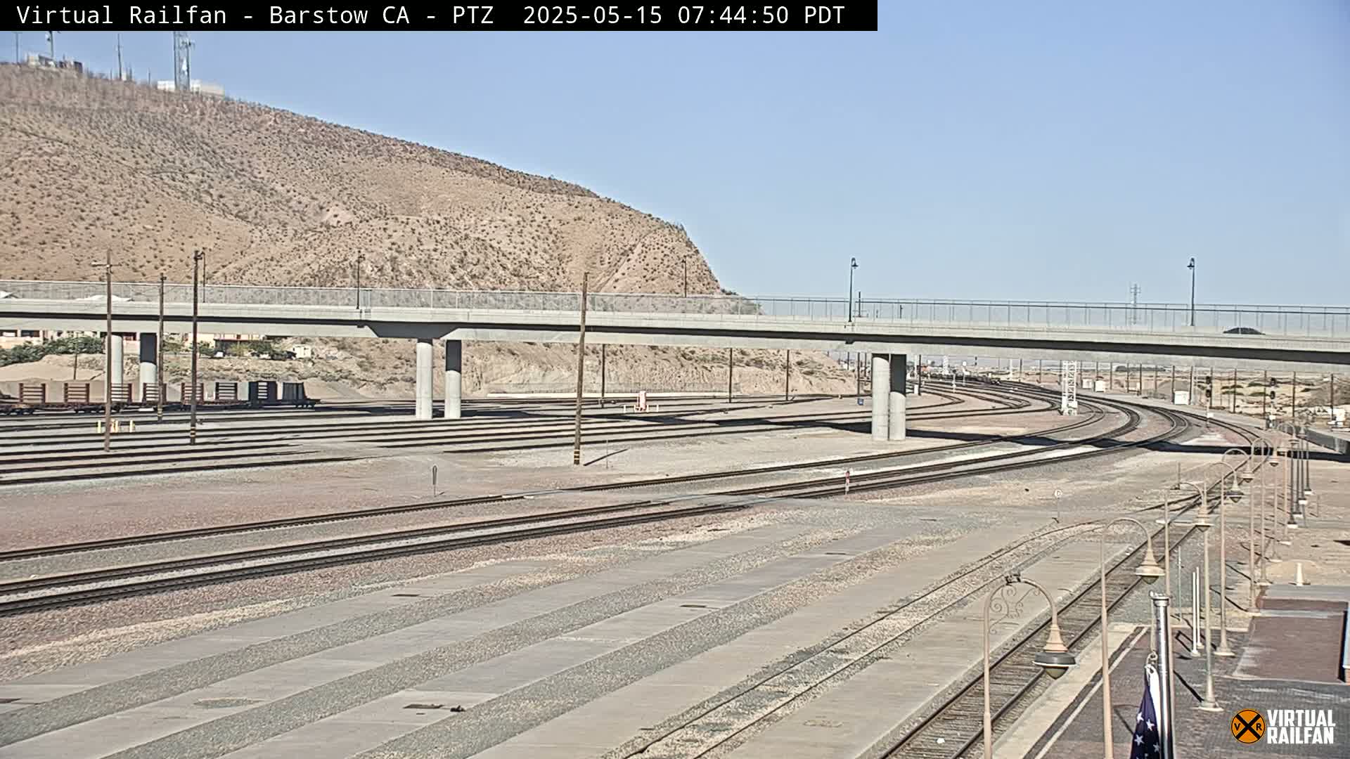 A long view of train tracks and a roadway overpass on a sunny day, with a desert landscape and hills in the background.