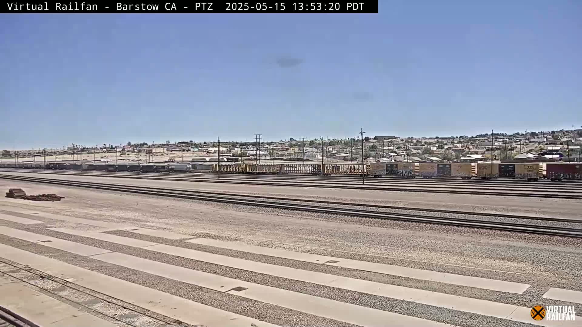 Under a clear sky, a long line of train cars sits on parallel railroad tracks in a large, flat, gravel-covered yard next to a residential area.