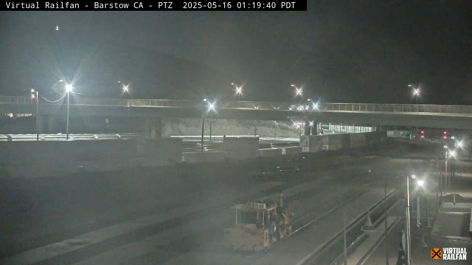 A nighttime view of a roadway overpass and railway yard, illuminated by streetlights.