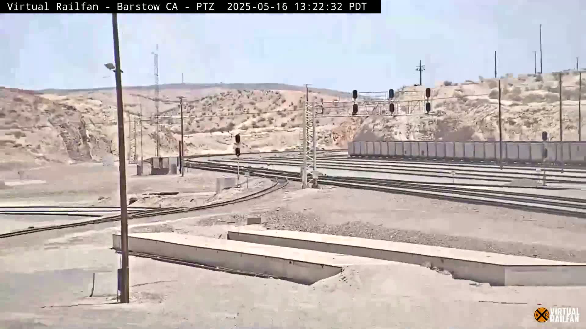 A sunny, desert landscape features a railway yard with multiple tracks, switching mechanisms, and a stationary freight train.