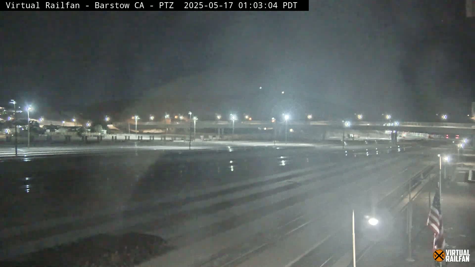 A nighttime view of a wet, empty roadway and train tracks under a dark sky illuminated by streetlights.