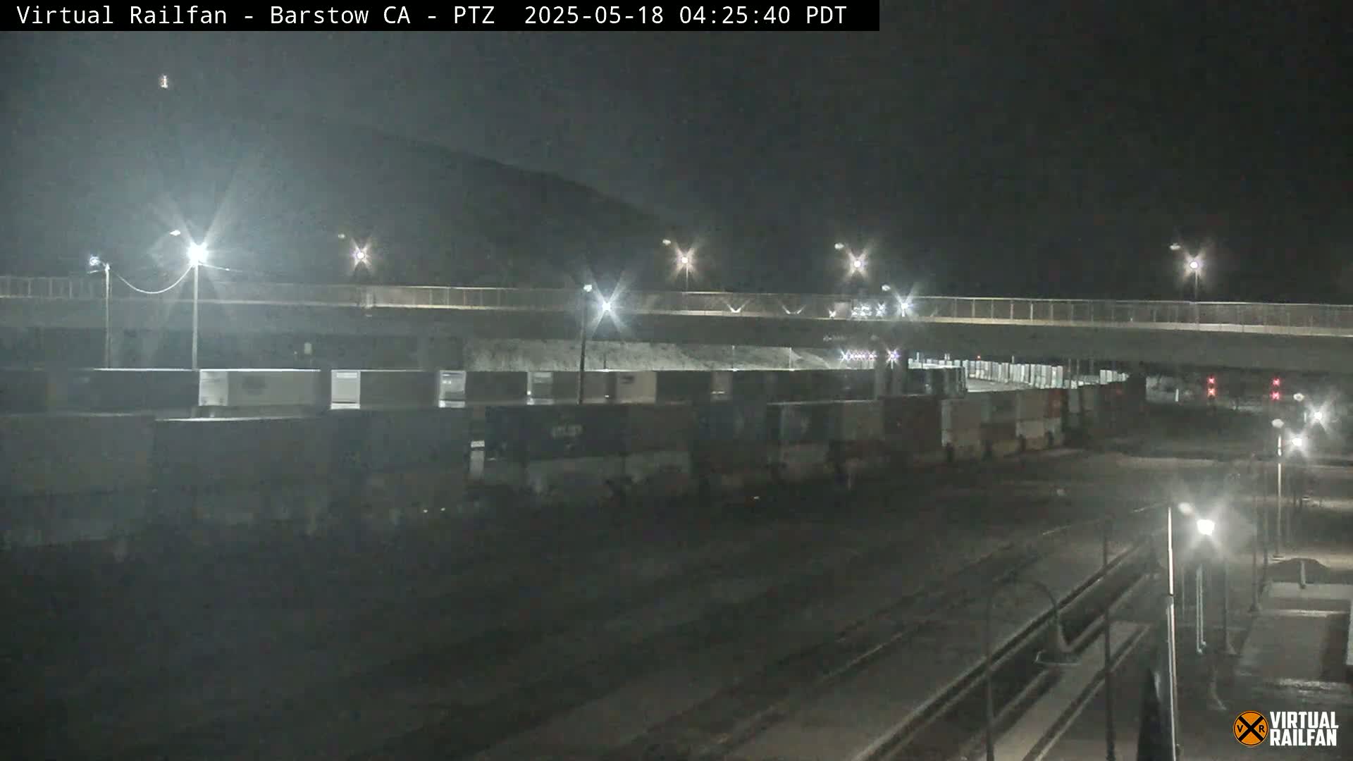 A nighttime view of a train yard with several boxcars under a bridge, illuminated by streetlights.