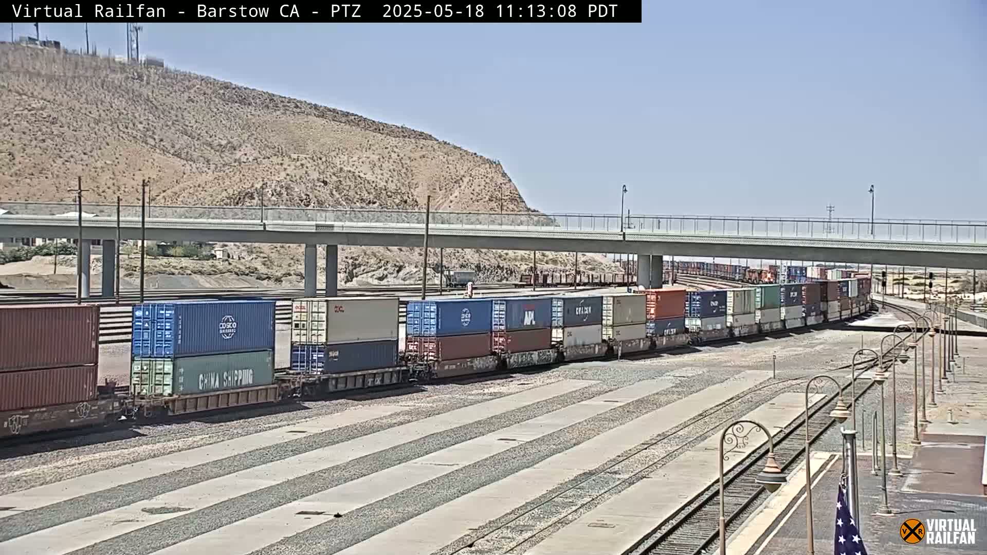 Under a clear sky, a long freight train carrying numerous shipping containers sits on the tracks near a highway overpass and a desert hillside.