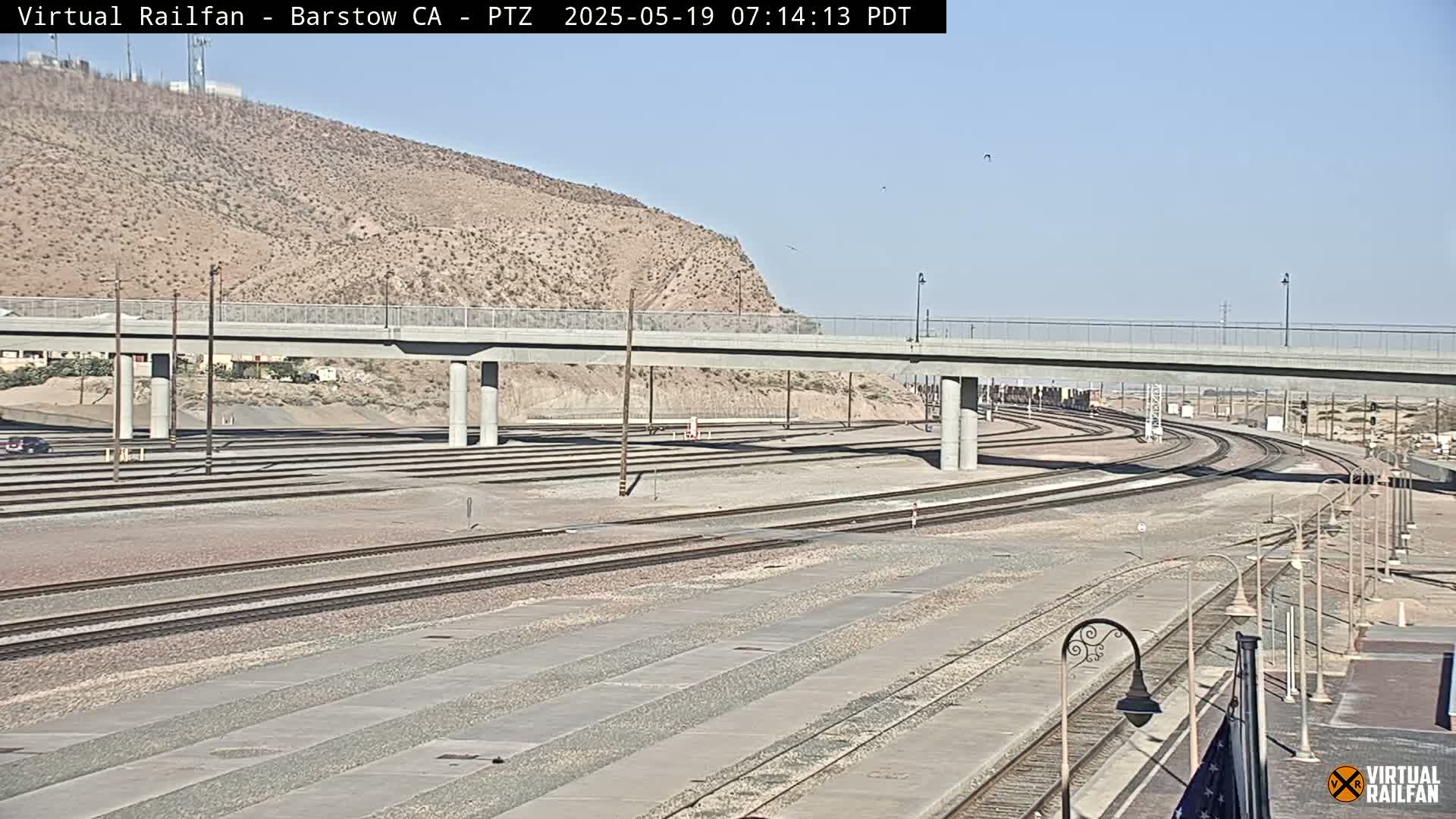 A wide shot of a railroad yard under a clear sky shows multiple train tracks, a road overpass, and a barren, rocky hill in the background.