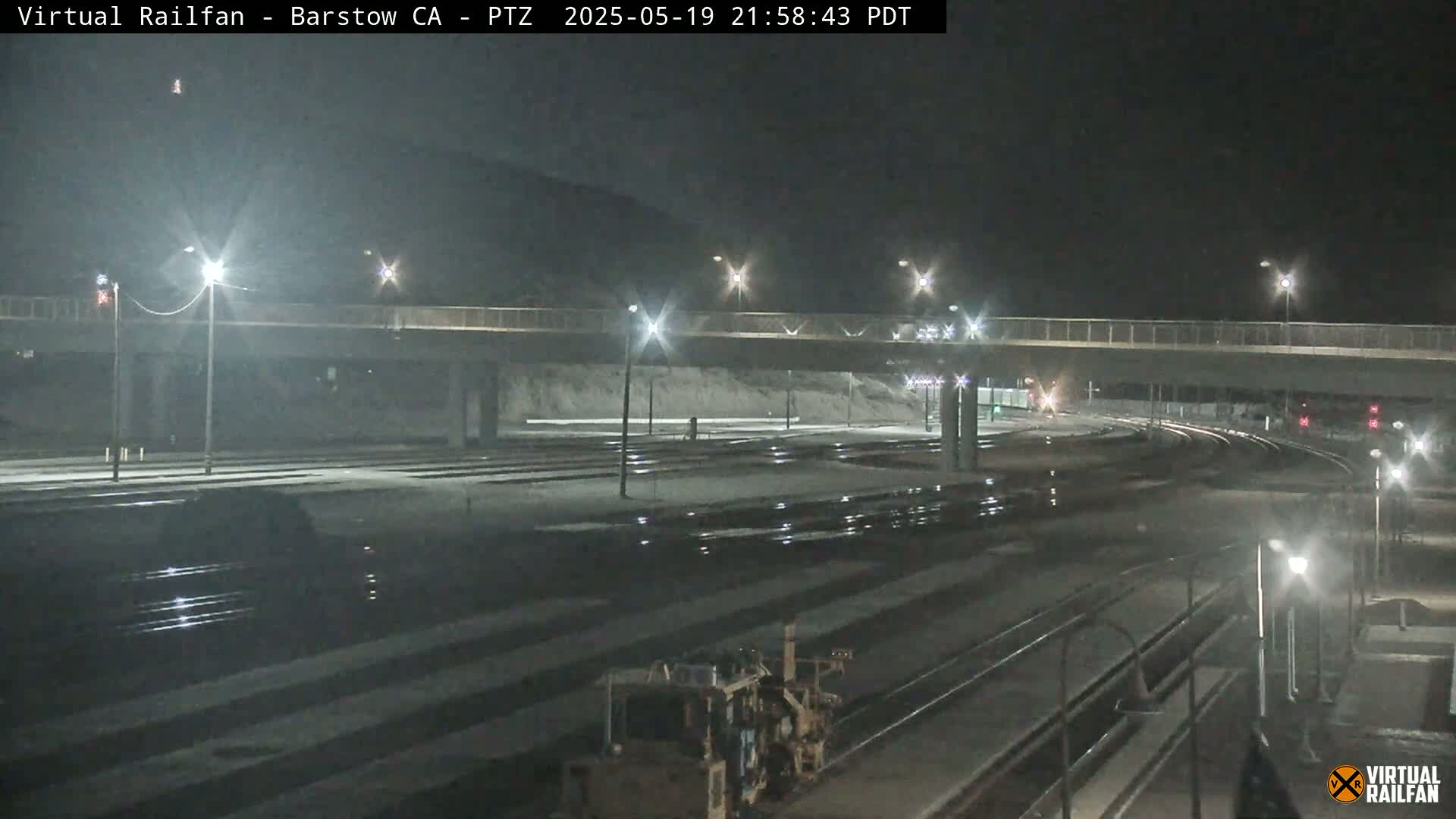 A nighttime view of a train yard with a road overpass, lit by streetlights, under a dark sky.
