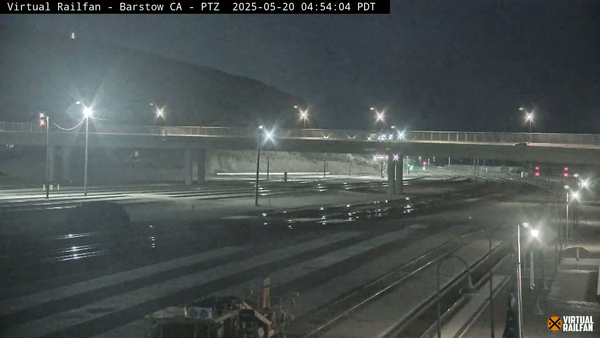 A nighttime view of a railroad yard with several sets of tracks, illuminated by streetlights, under a bridge and near a hill.