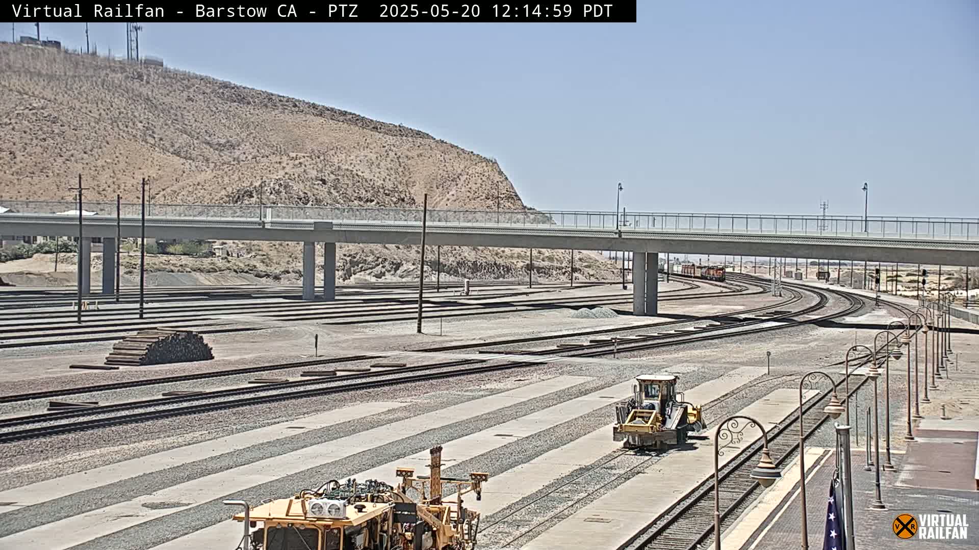 A sunny day reveals a wide view of railroad tracks, construction equipment, and a road bridge over the tracks, all set against a backdrop of a dry, rocky hill.