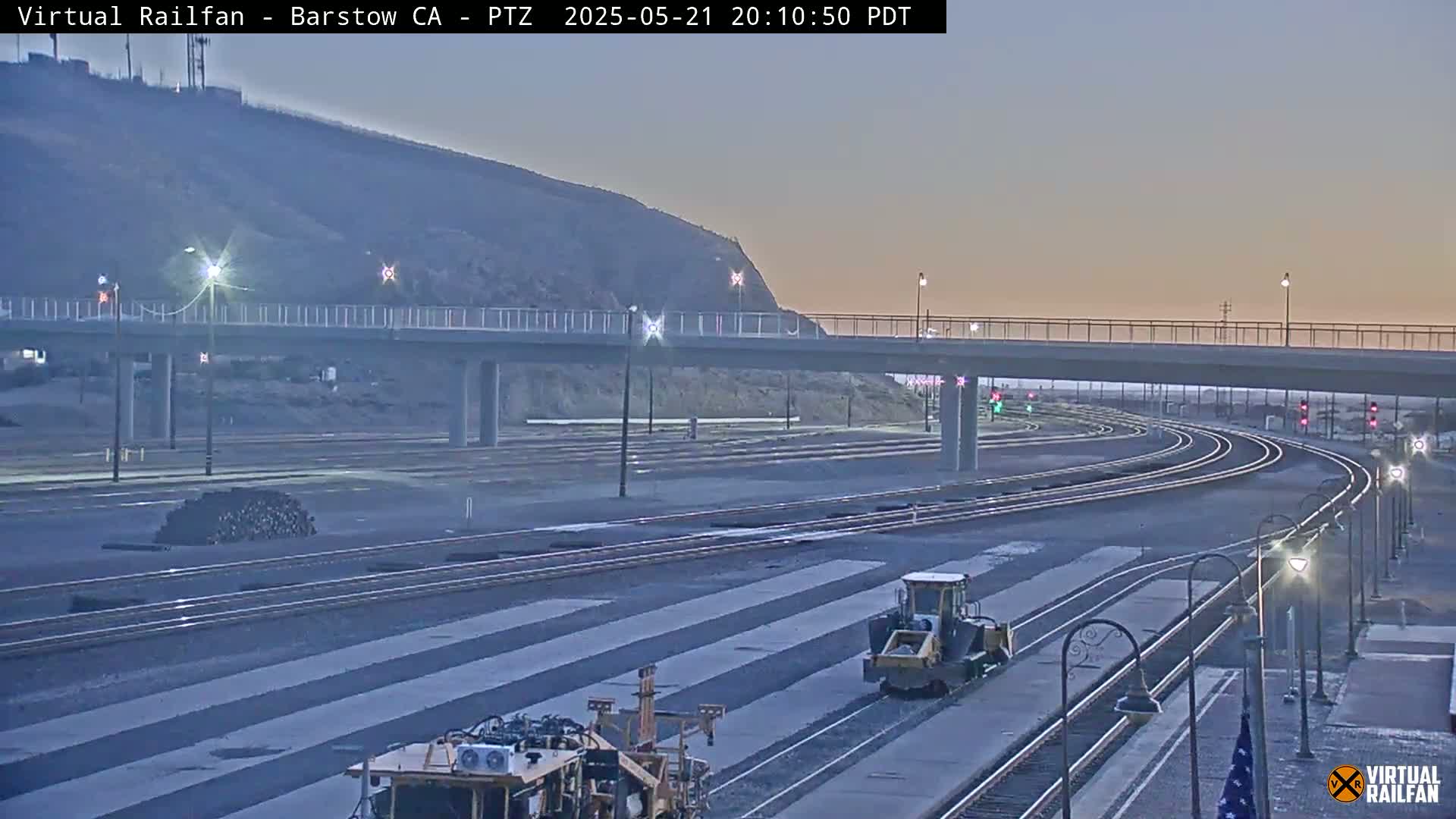 Under a twilight sky, two pieces of heavy machinery work on a railway yard near an overpass.