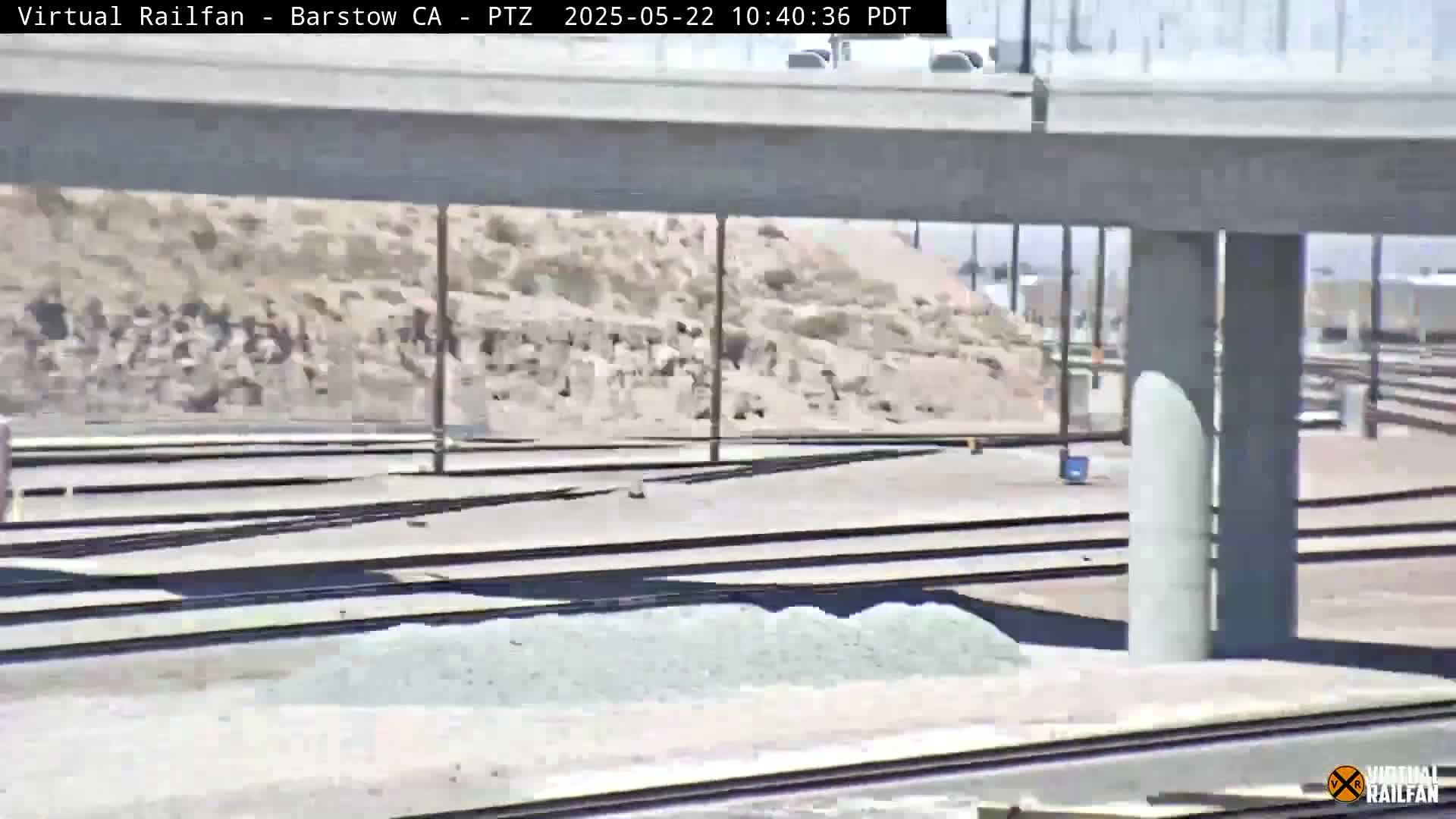 A view of railroad tracks under a highway overpass on a sunny day, with a pile of gravel nearby.