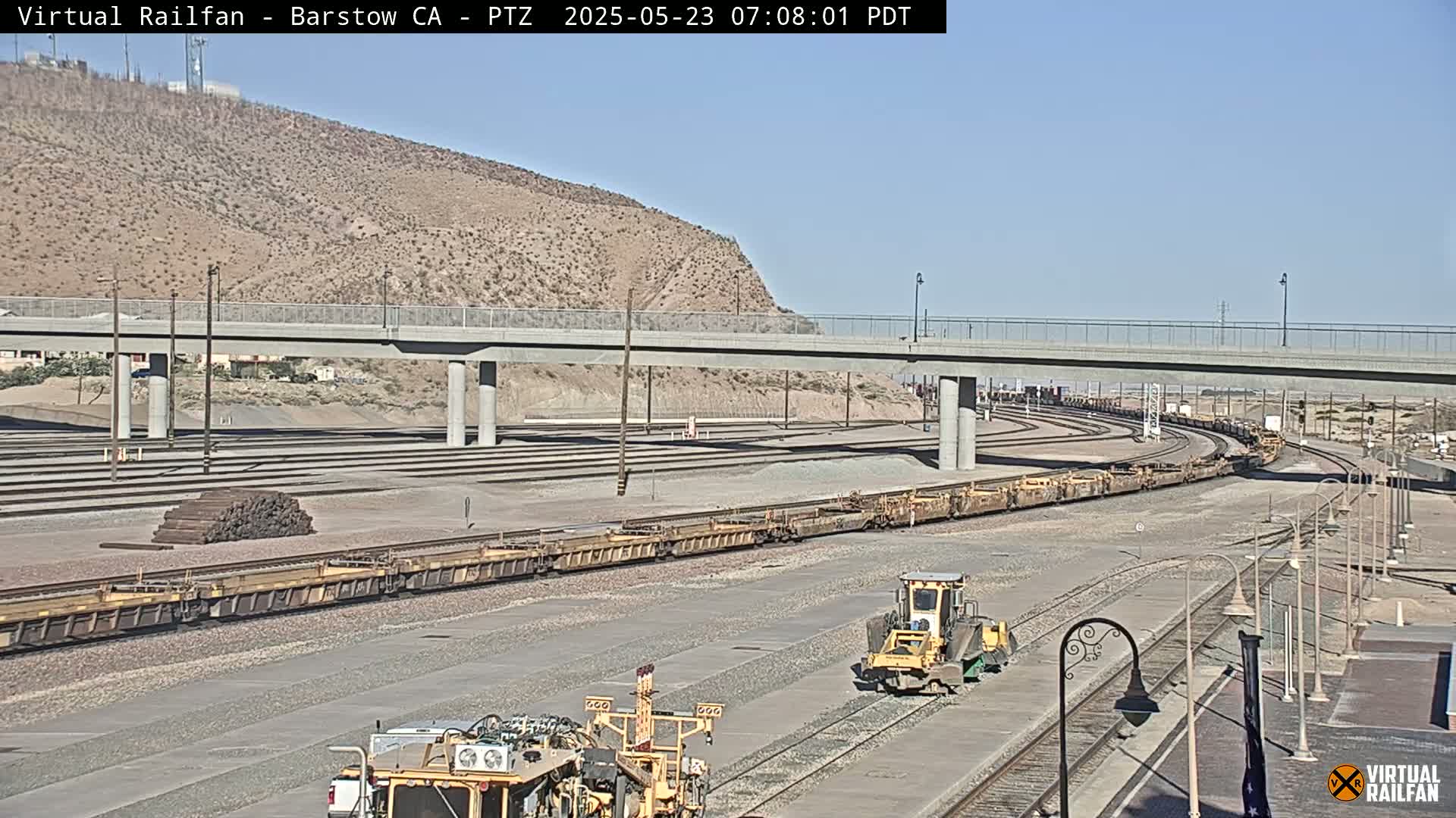 A long freight train is passing under a bridge on a sunny day, with railway maintenance equipment visible nearby.