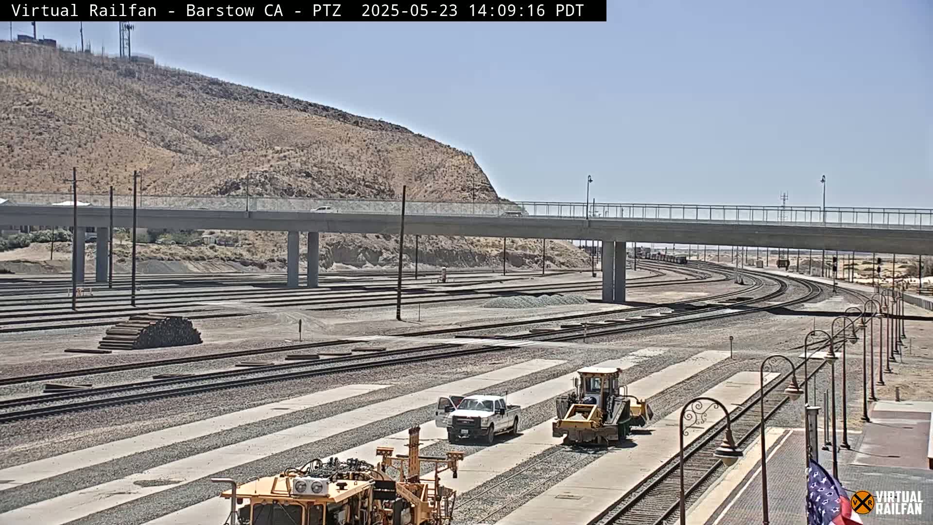 Under a clear sky, a wide expanse of railroad tracks and switching yards are shown, with several pieces of construction or maintenance equipment present near a road overpass.