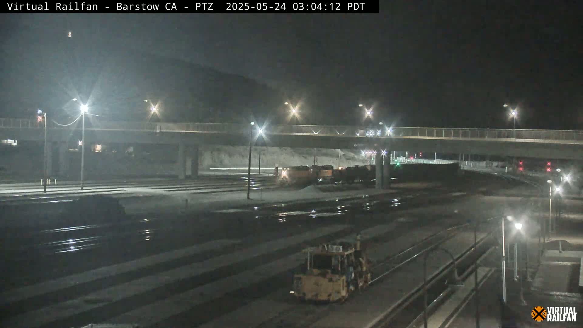 A nighttime view of a railway yard with a train and a piece of heavy machinery, under partly cloudy skies.