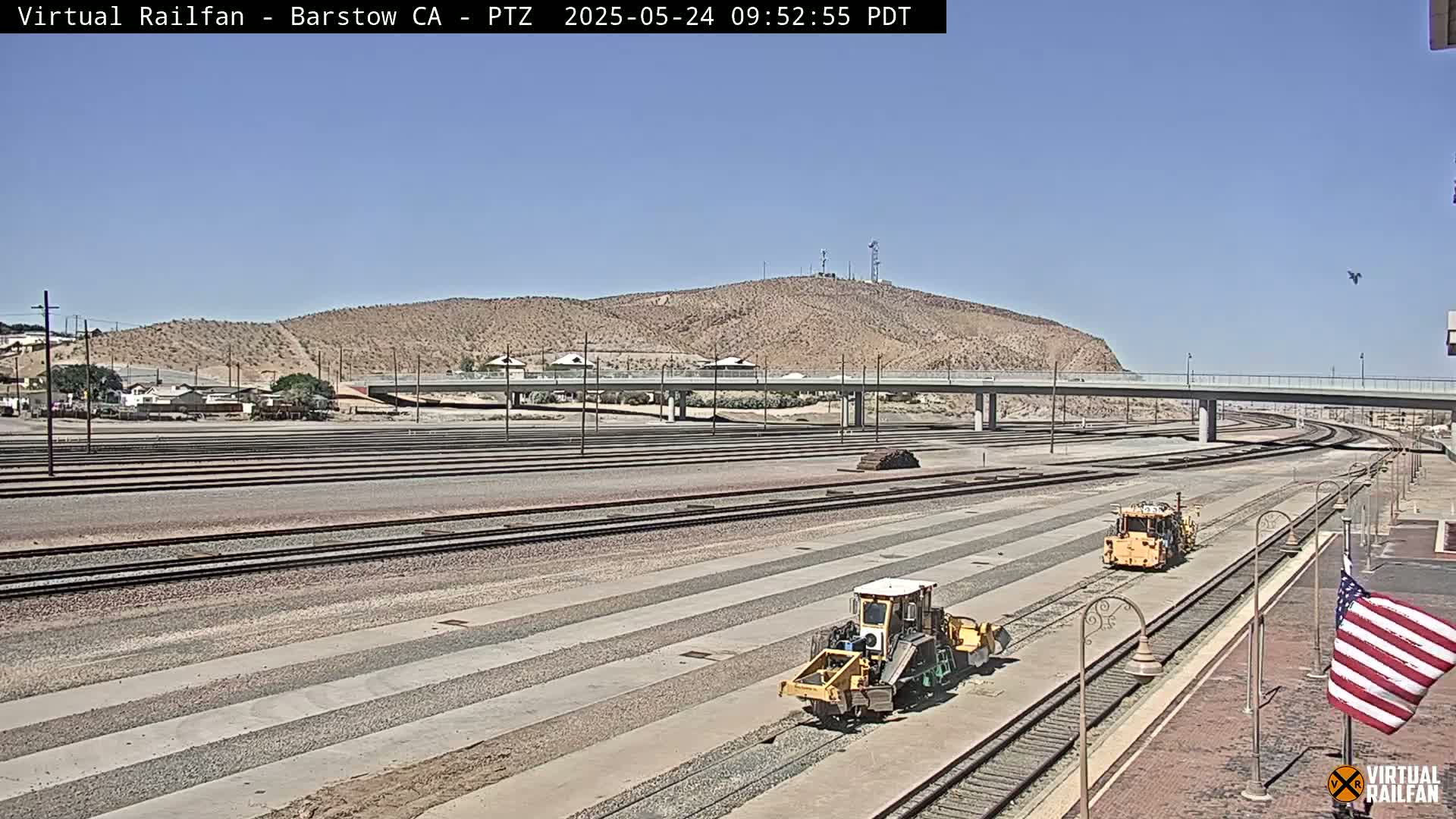 Under a clear sky, two pieces of yellow railway maintenance equipment sit on multiple parallel railway tracks that stretch toward a road overpass and a desert hillside.