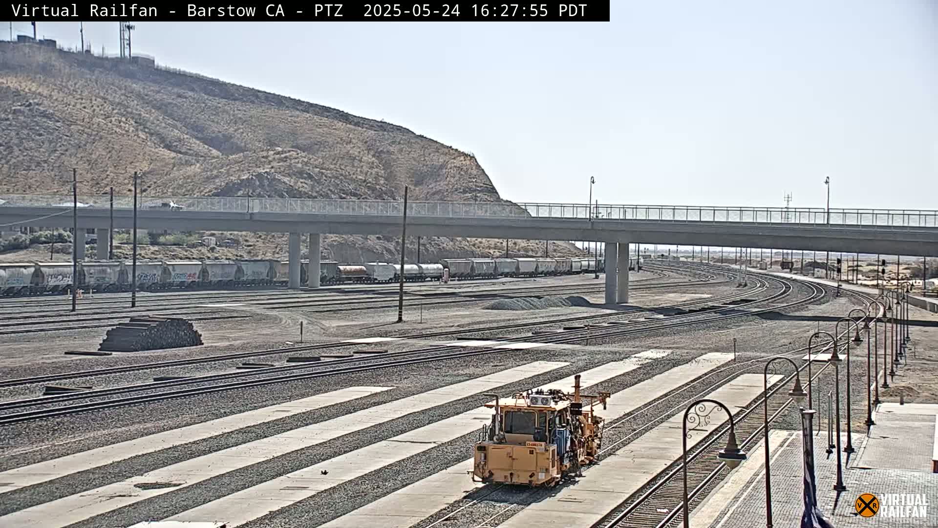 A yellow rail maintenance vehicle is positioned on a set of train tracks in a sunny, outdoor rail yard, with a parked freight train visible in the background.