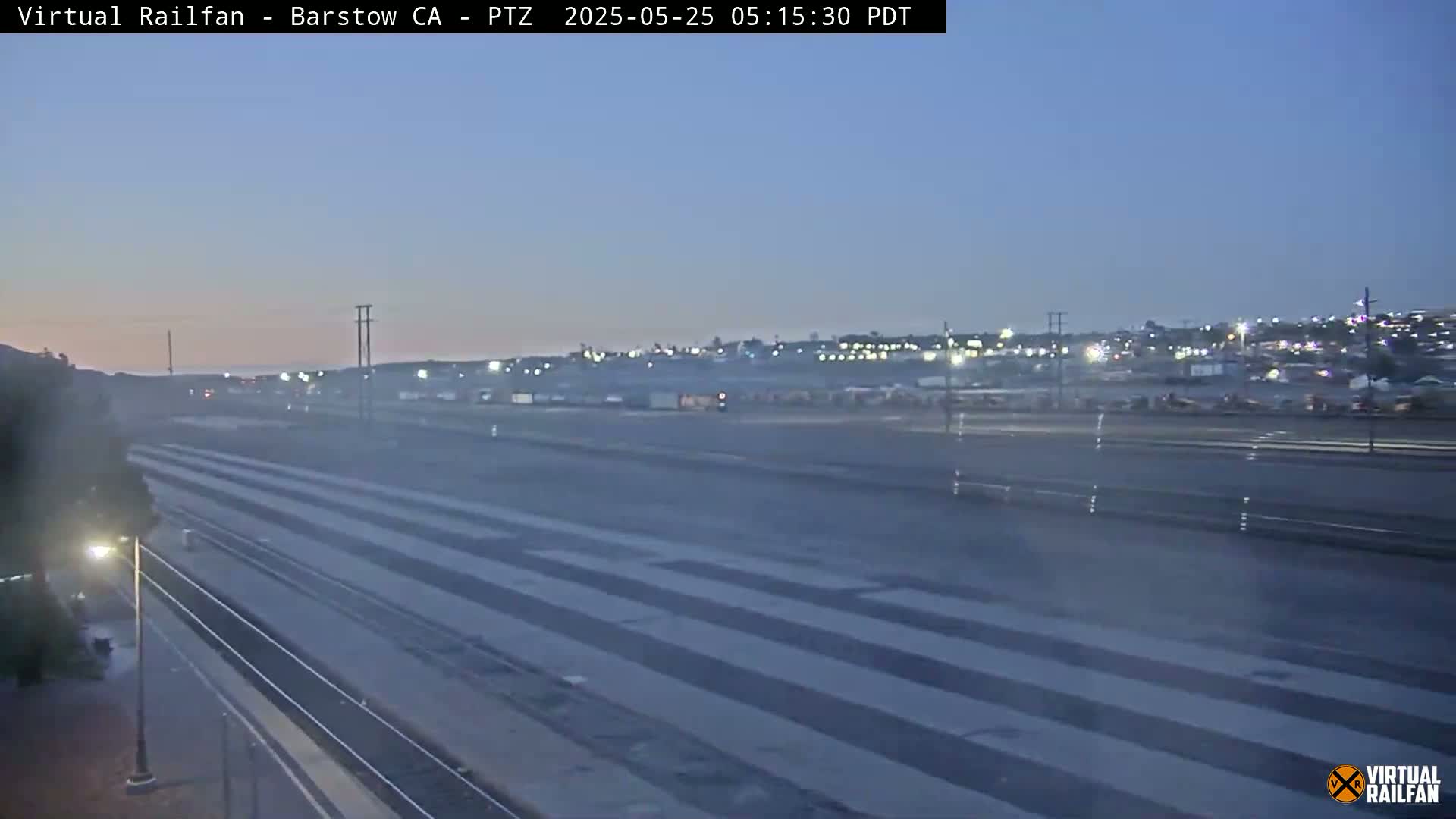 A twilight view of a multi-lane roadway and railway tracks, with a hazy or foggy atmosphere and distant city lights visible on the horizon.