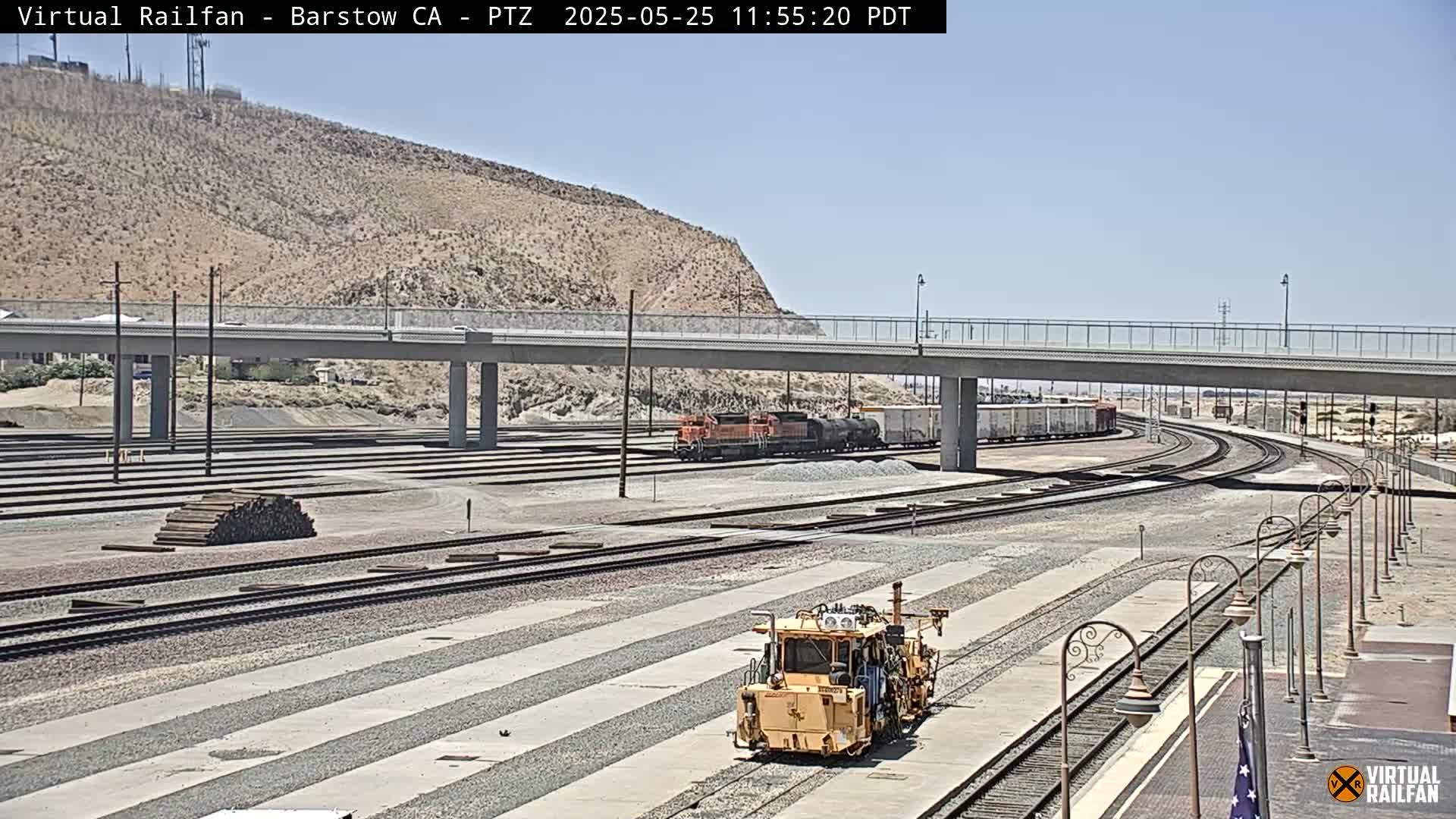 A yellow rail maintenance vehicle sits in a rail yard on a sunny day, with a train in the background and a bridge overhead.